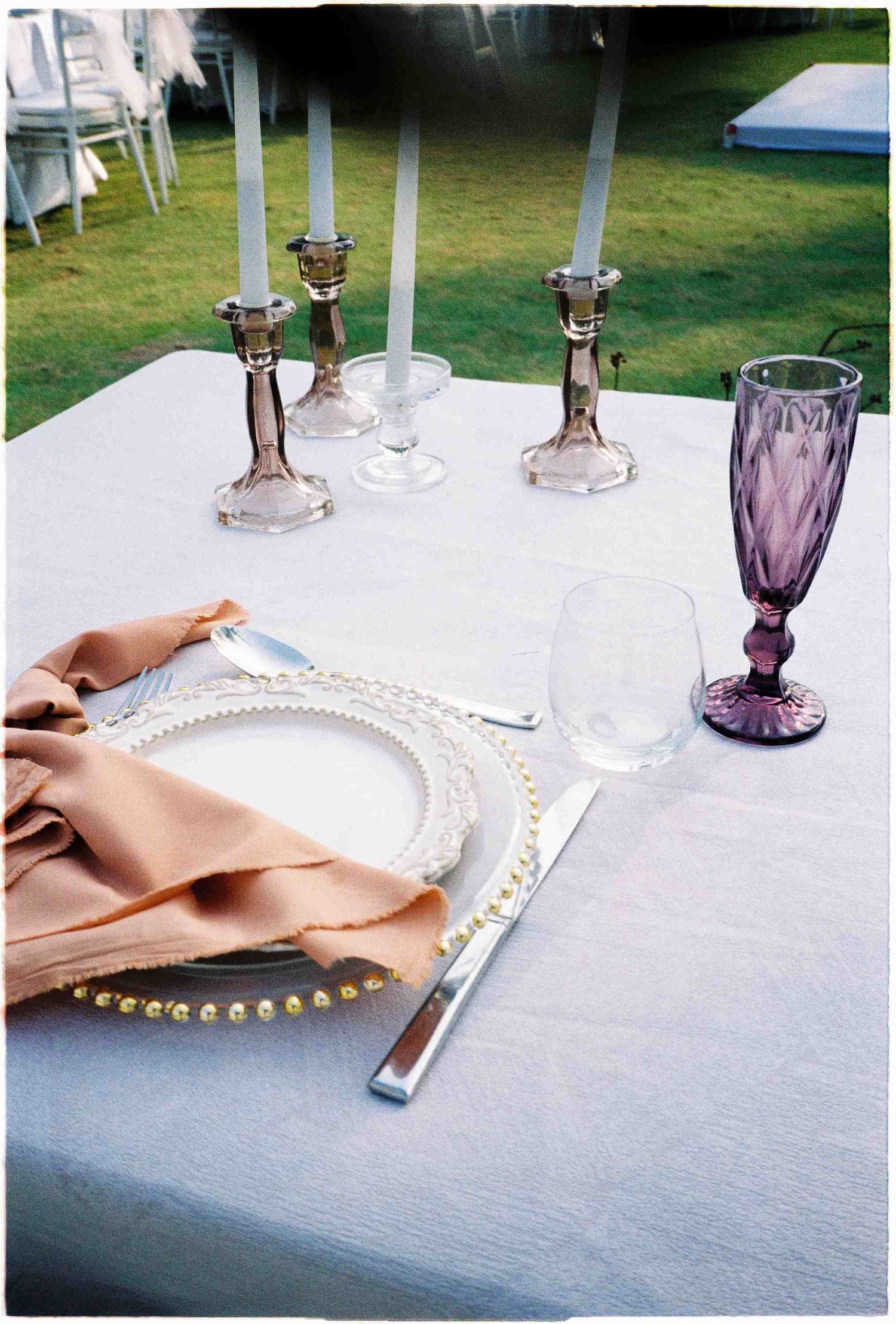 Elegant outdoor table setting with pearl-rimmed plate, peach napkin, silver candlesticks, and purple goblet on white linen.