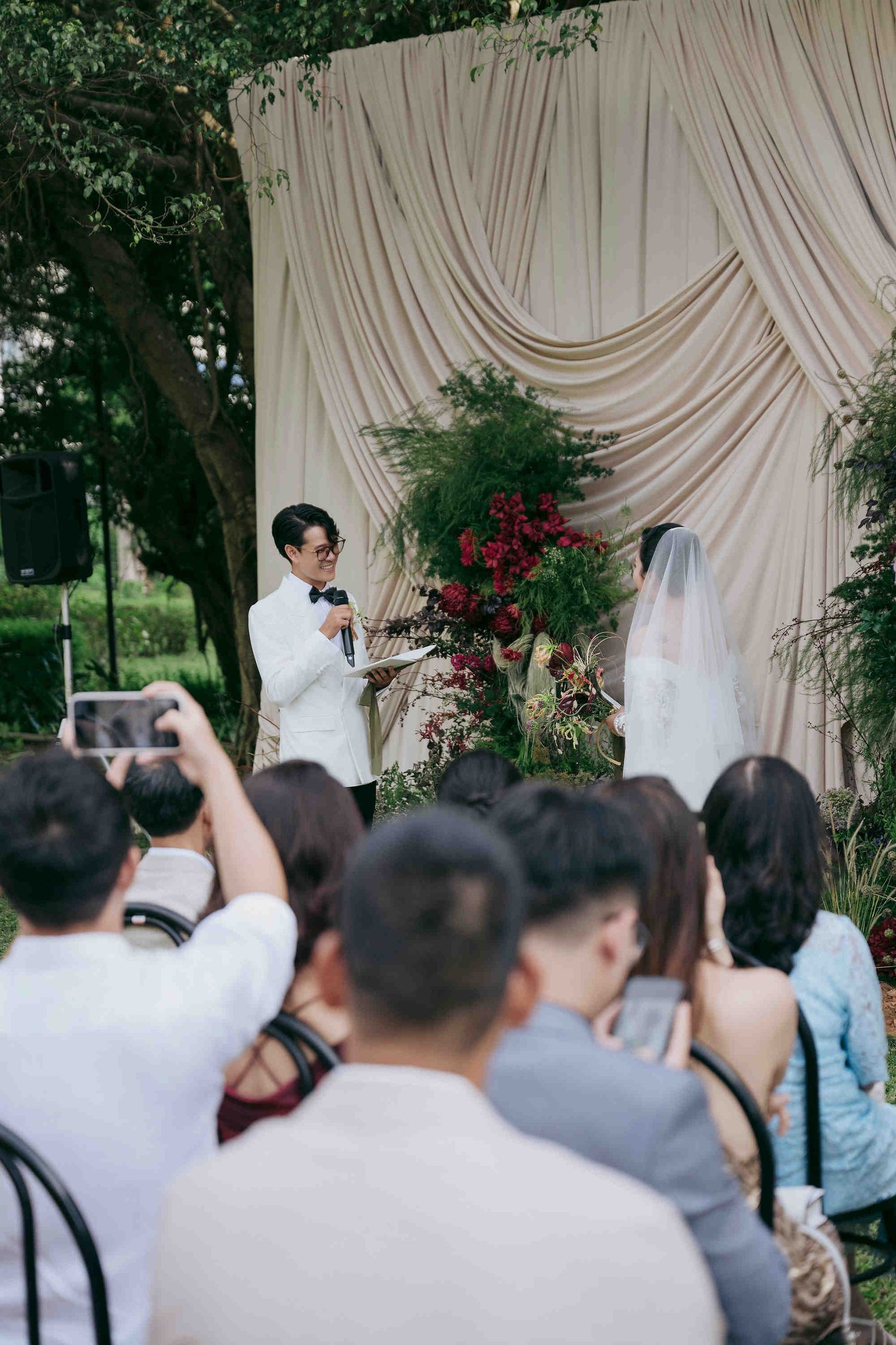 Groom in white tuxedo reciting vows to veiled bride at outdoor wedding ceremony with floral backdrop.