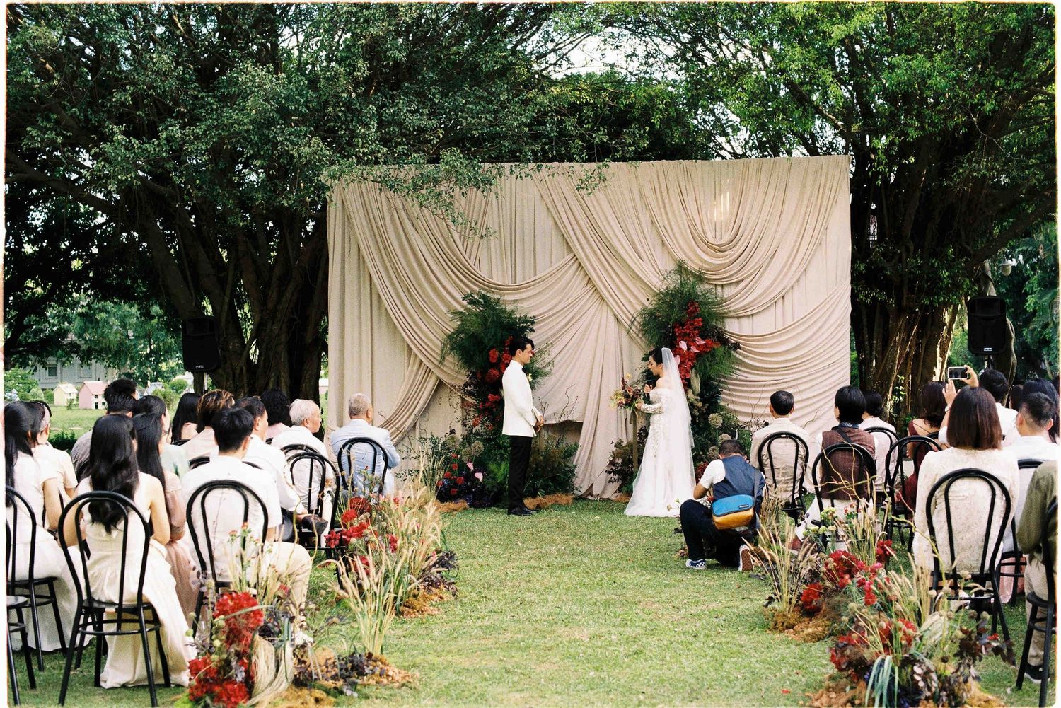 Outdoor wedding ceremony on green lawn with draped fabric backdrop, floral arrangements, and guests seated in black chairs.