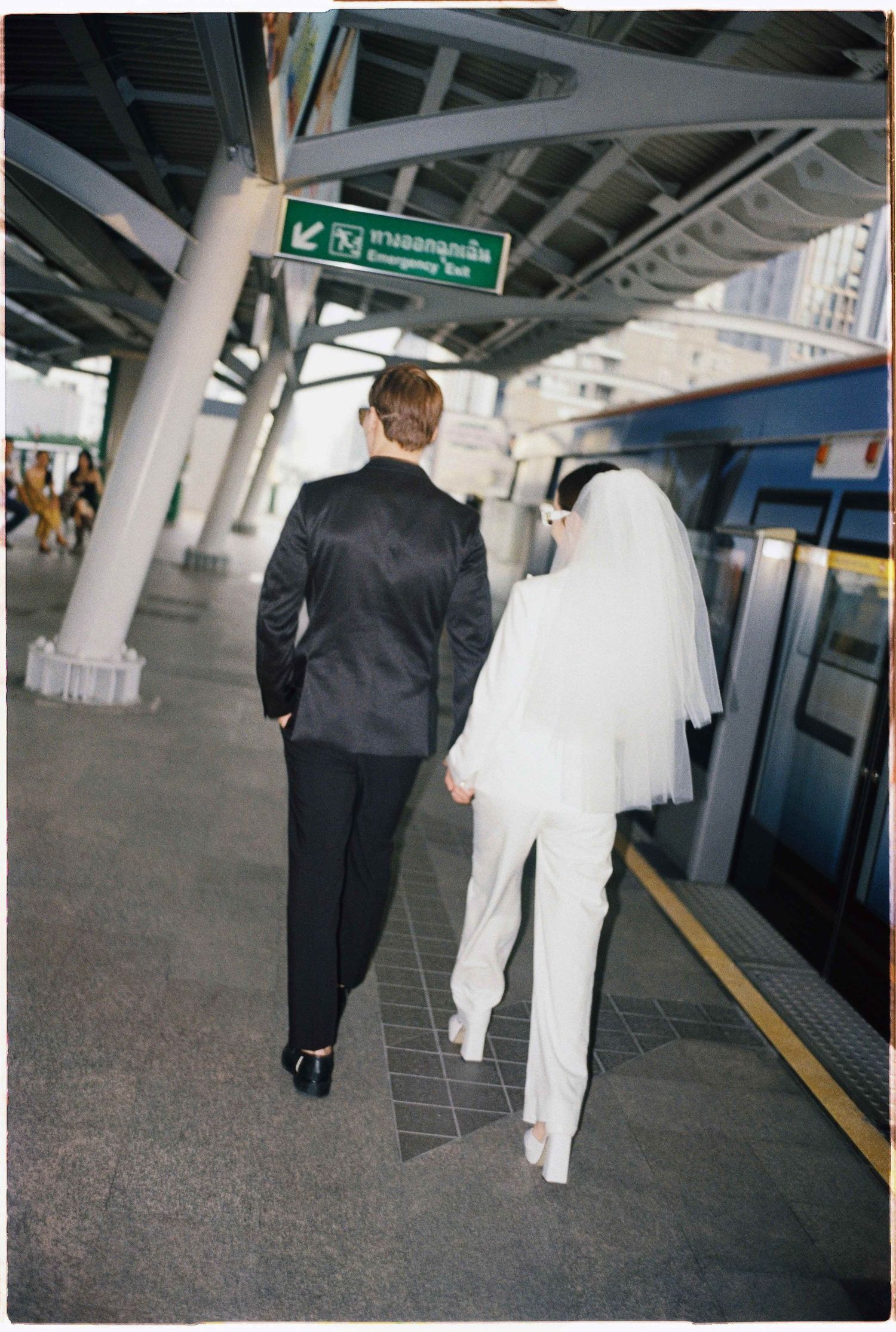 Saigon pre-wedding: couple in black suit and white outfit with veil walk along a modern transit platform, by AnhVuWedding.