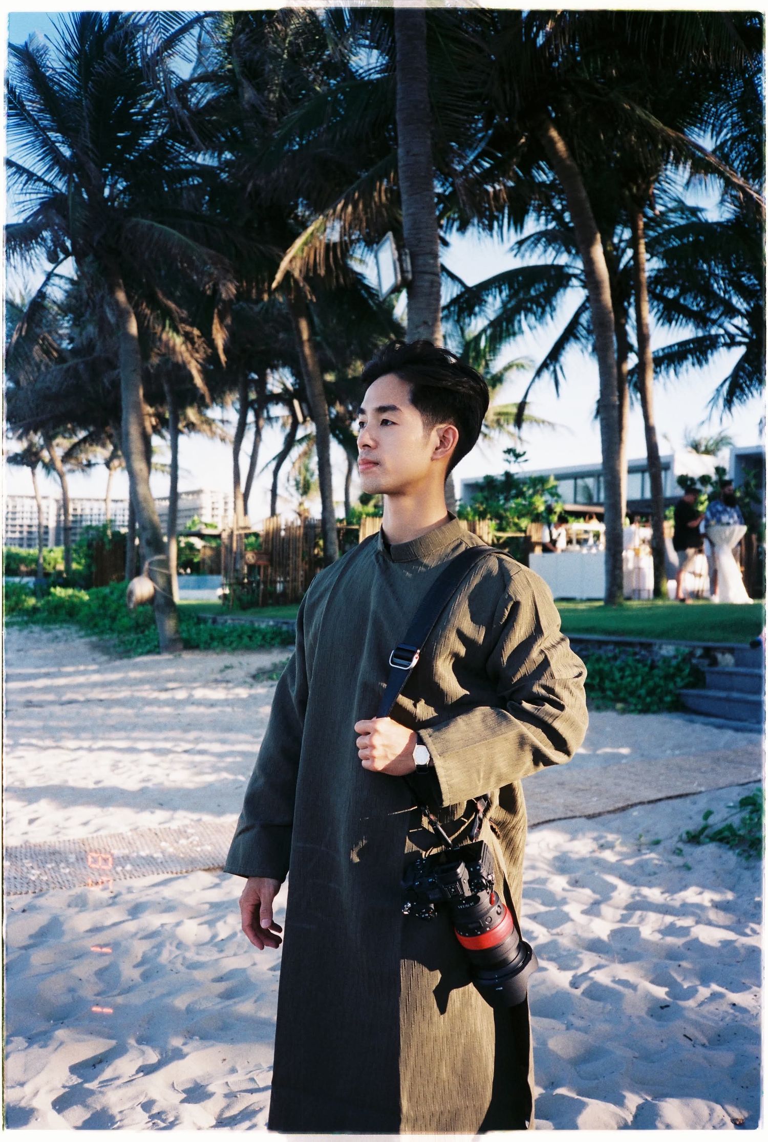 portrait photo: A person in a long dark coat stands on a beach path with palm trees and tropical scenery in the background, by AnhVuWedding.