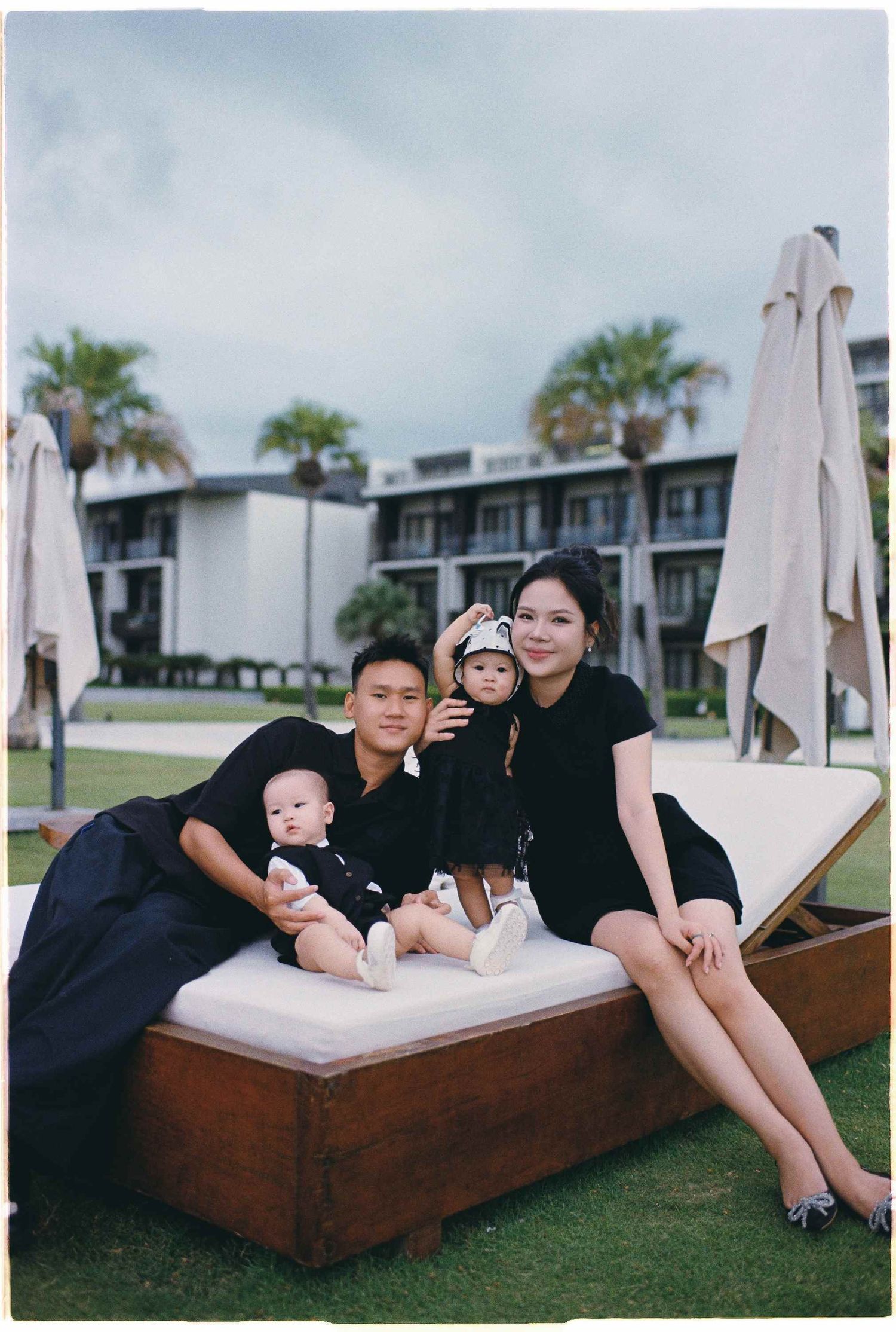Da Nang family: family dressed in black sits together on an outdoor daybed at a tropical resort, by AnhVuWedding.