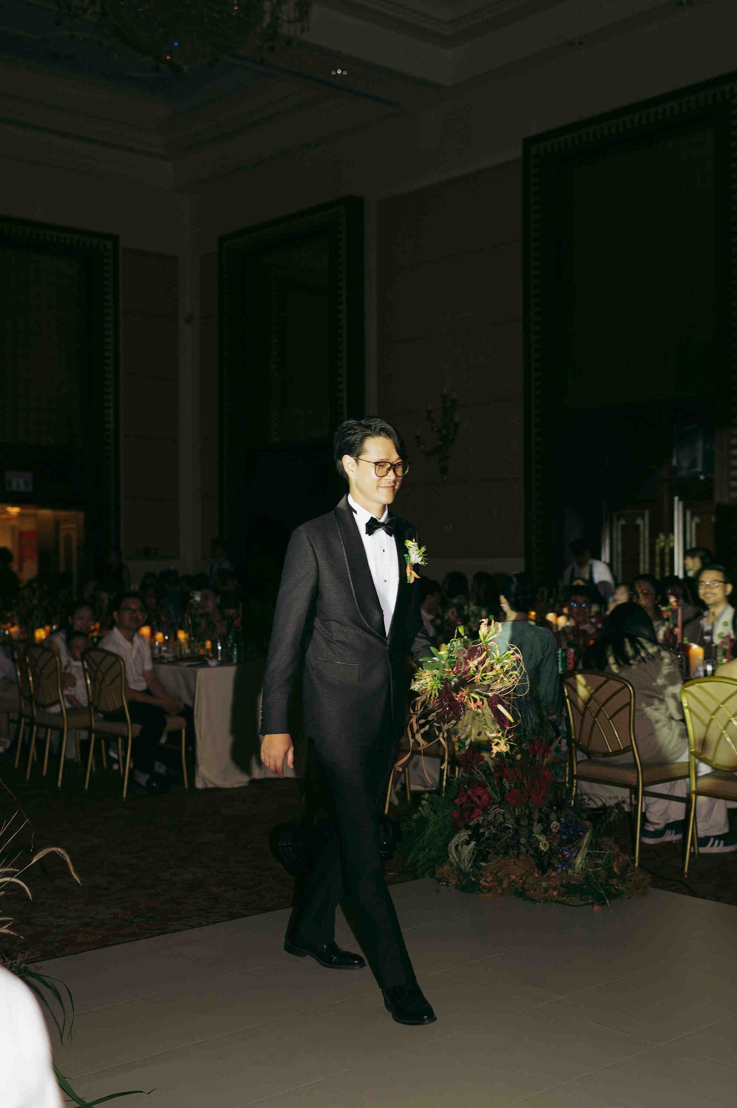 Groom in black tuxedo with bow tie and boutonniere walks at elegant wedding reception venue.