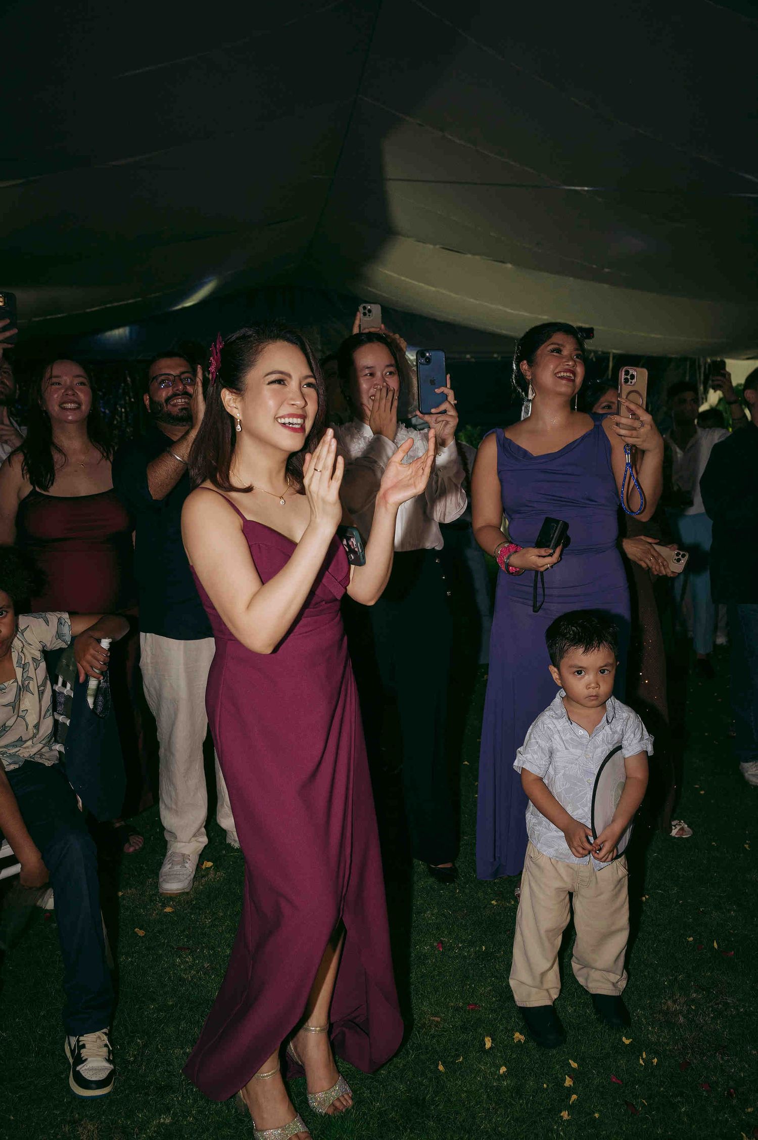 Guests in elegant attire clap and smile at a wedding reception, with a young boy standing nearby.