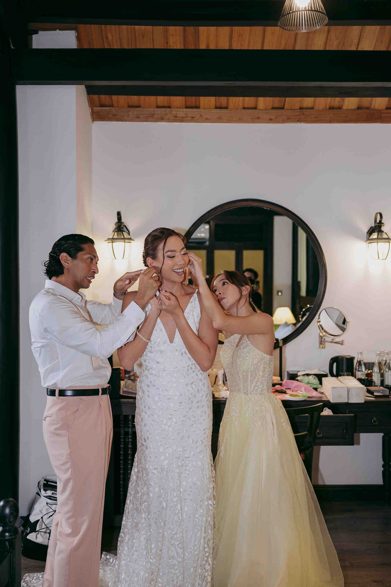 Bride in white lace gown gets earrings put on by two helpers in elegant dressing room before wedding ceremony.