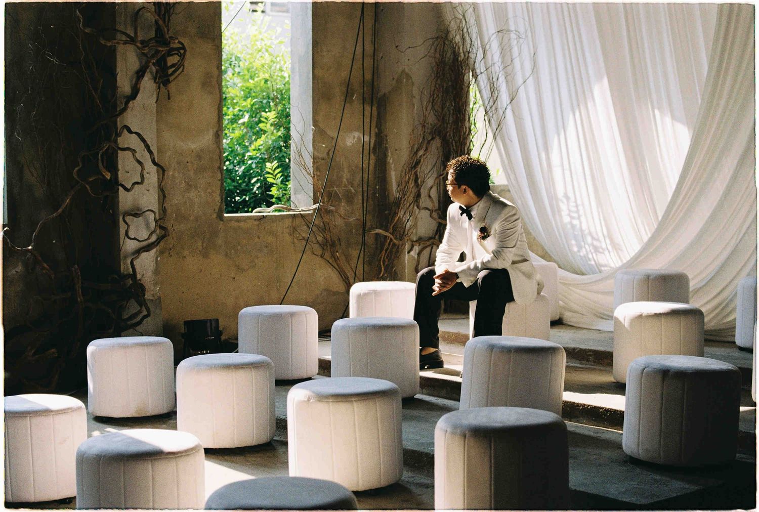 A person sits among white cylindrical ottomans in a rustic room with draped curtains and aged walls.