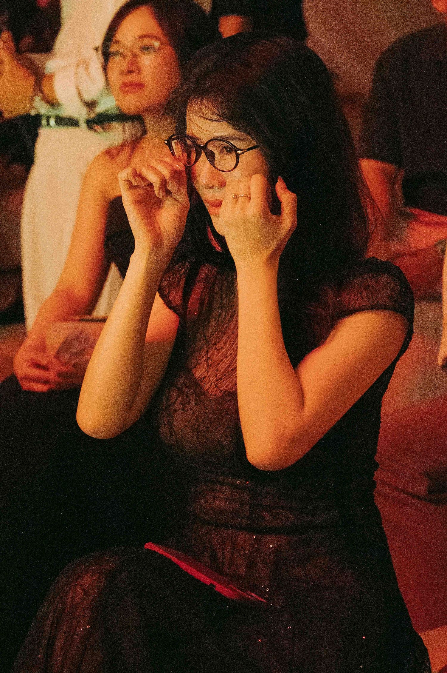 Woman in black lace dress adjusting round glasses while seated at an event, warm ambient lighting.