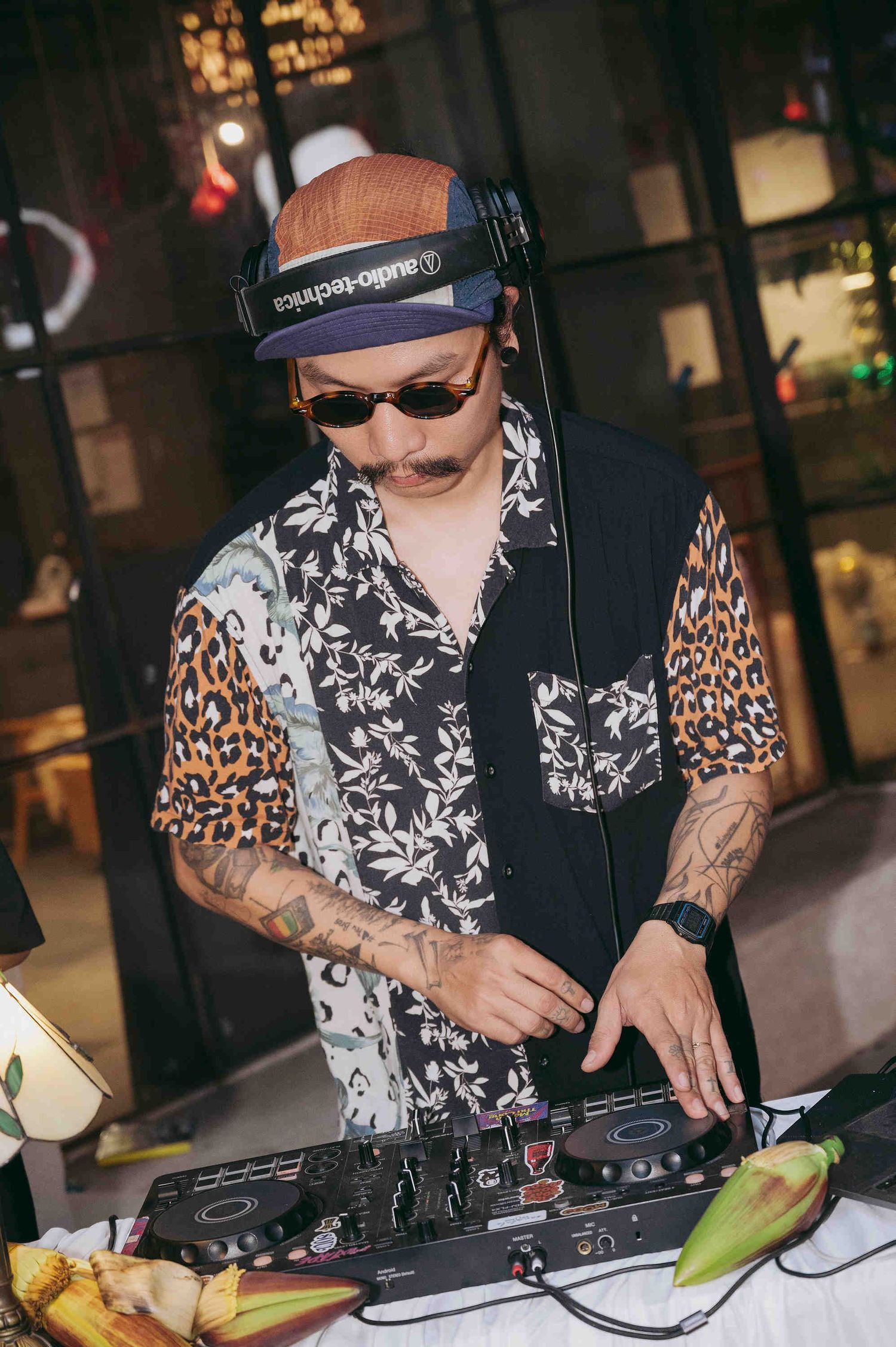 Tattooed DJ wearing headphones, leopard print shirt, and cap mixing music on DJ equipment at an outdoor event.