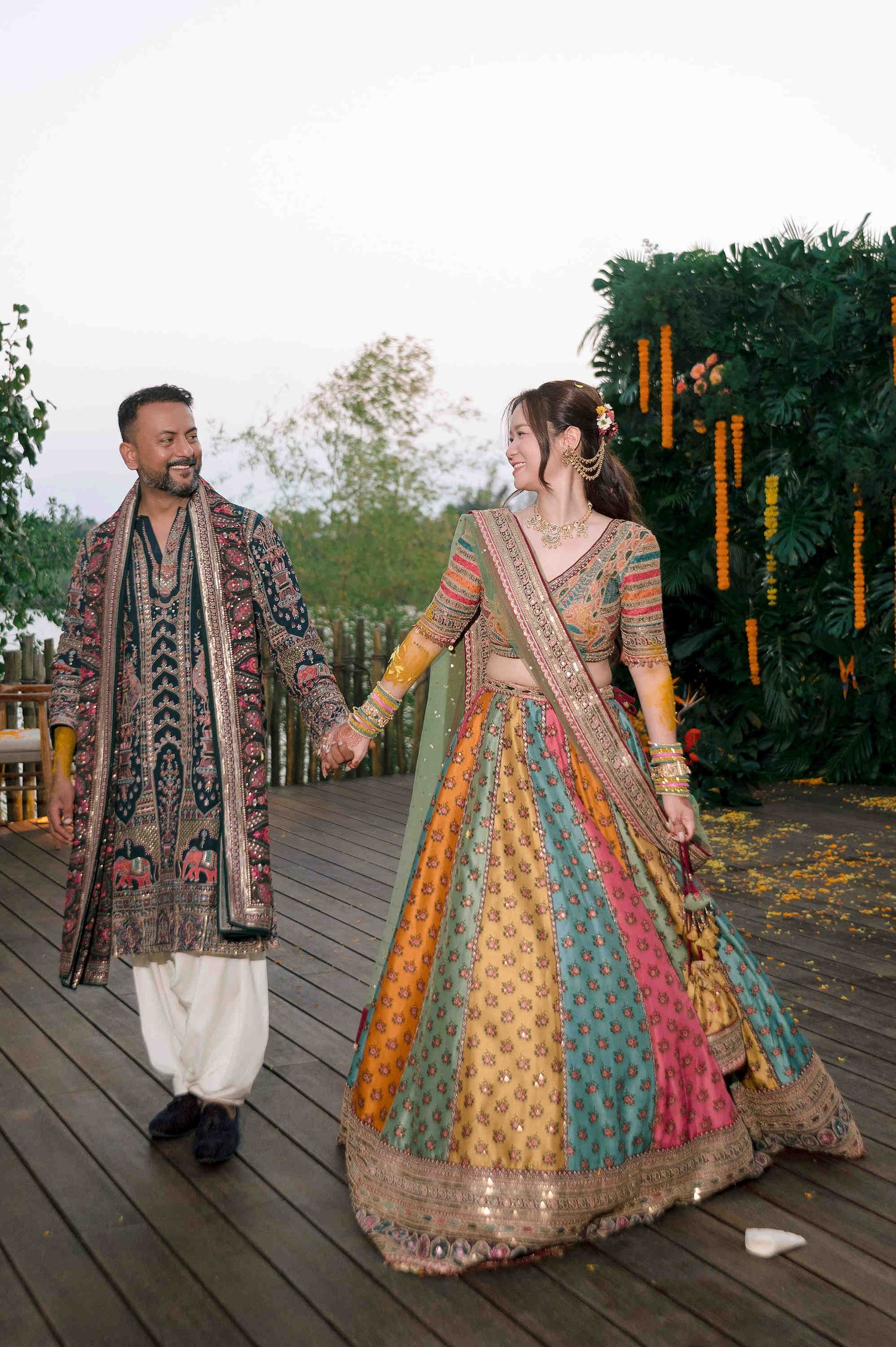Bride and groom in colorful Indian wedding outfits walking hand in hand on a decorated wooden deck.