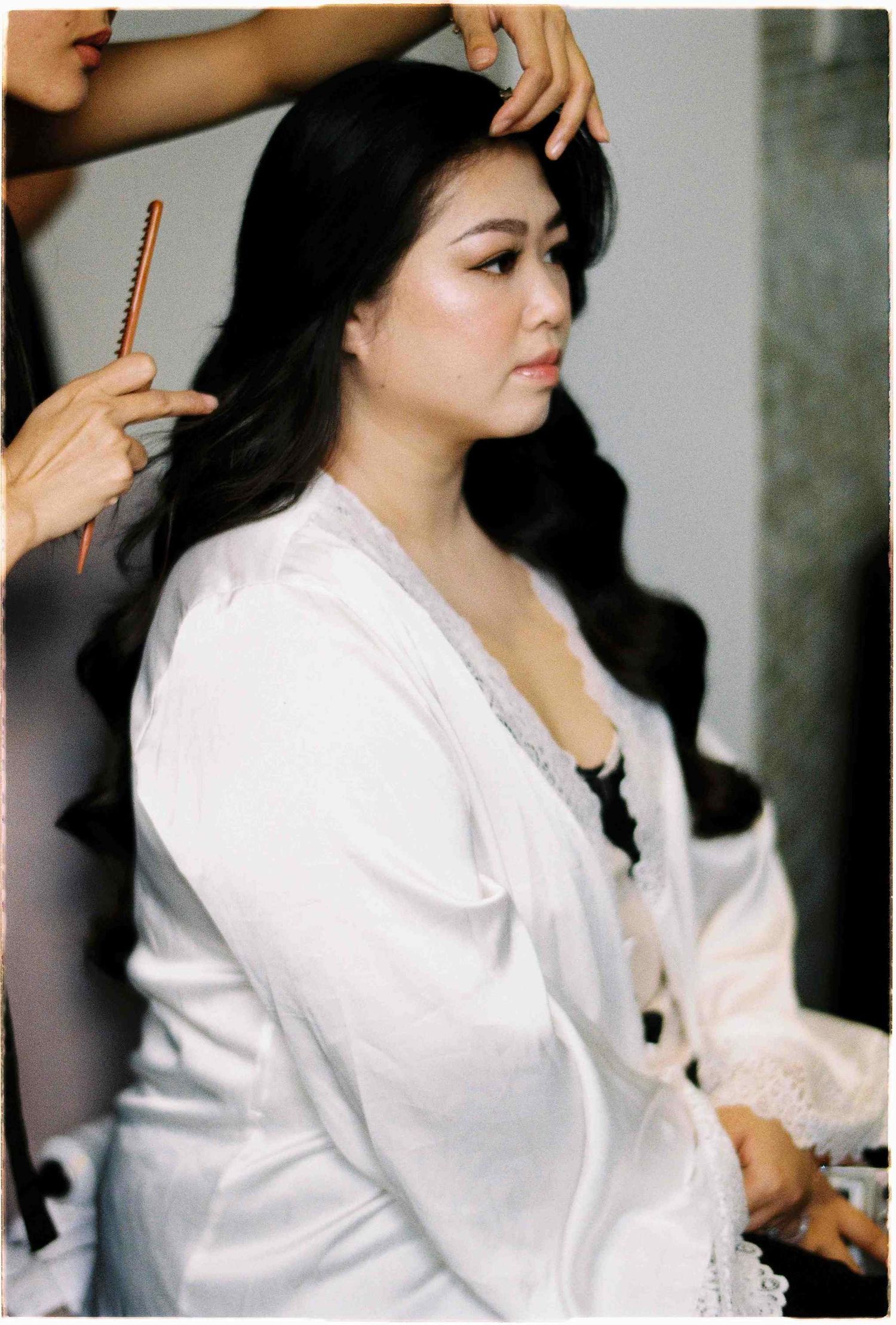 A bride in a white robe gets her long dark hair styled with a comb during wedding preparations.