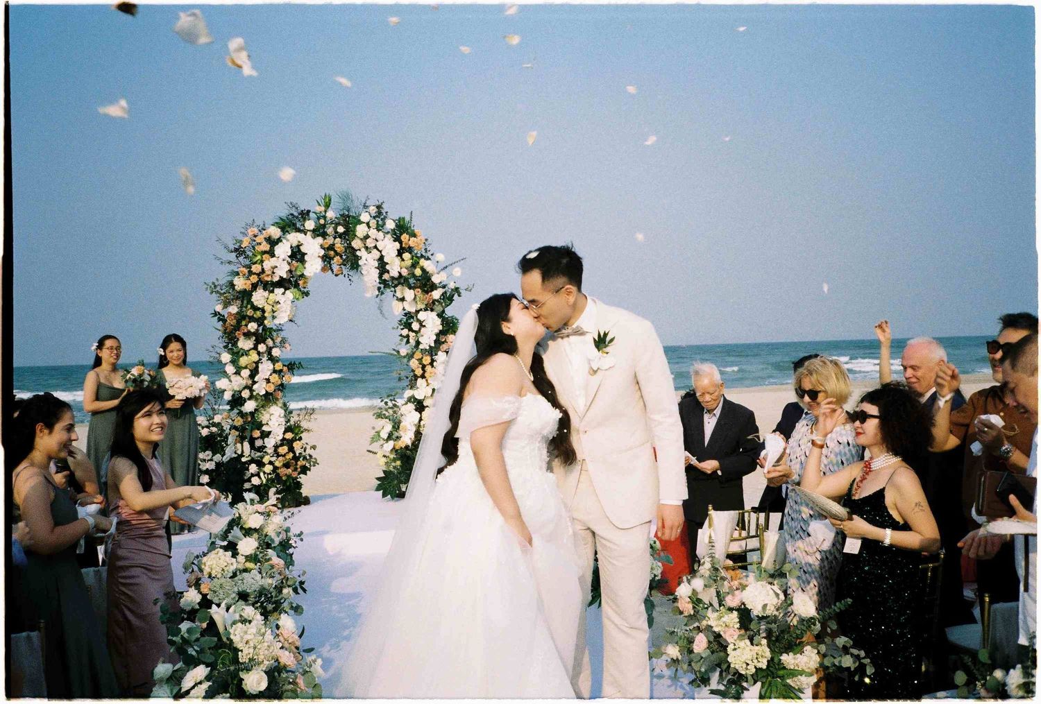 Bride and groom share first kiss at beach wedding ceremony with floral arch and flower petals.