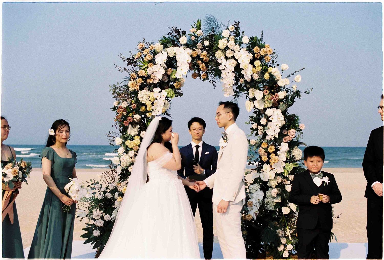 Couple exchanges vows at a beach wedding ceremony under a floral arch with bridesmaids and guests.