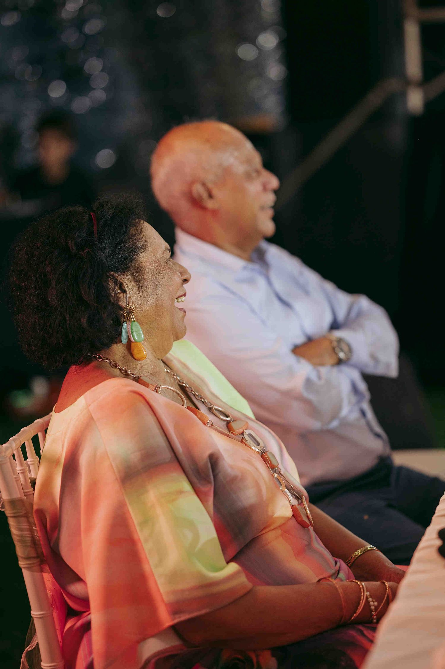 Elderly couple seated at an event, woman in colorful sari with turquoise earrings smiling, man in blue shirt with arms crossed.