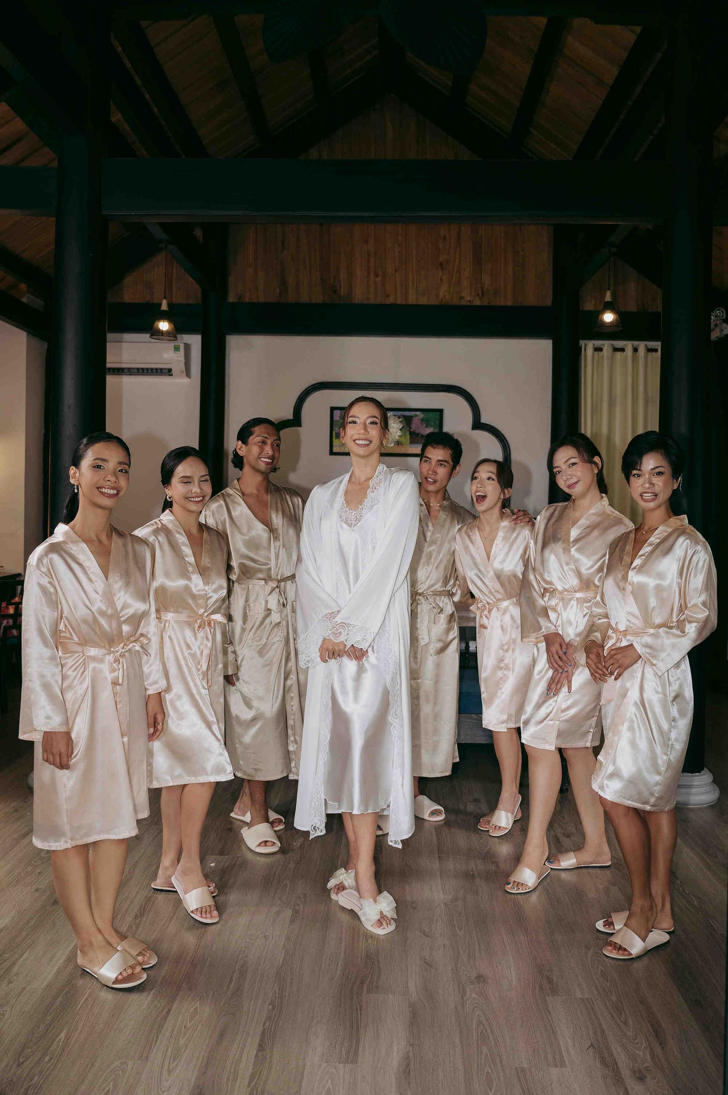 Bride in white robe poses with bridesmaids in champagne satin robes and slippers inside elegant wooden venue.