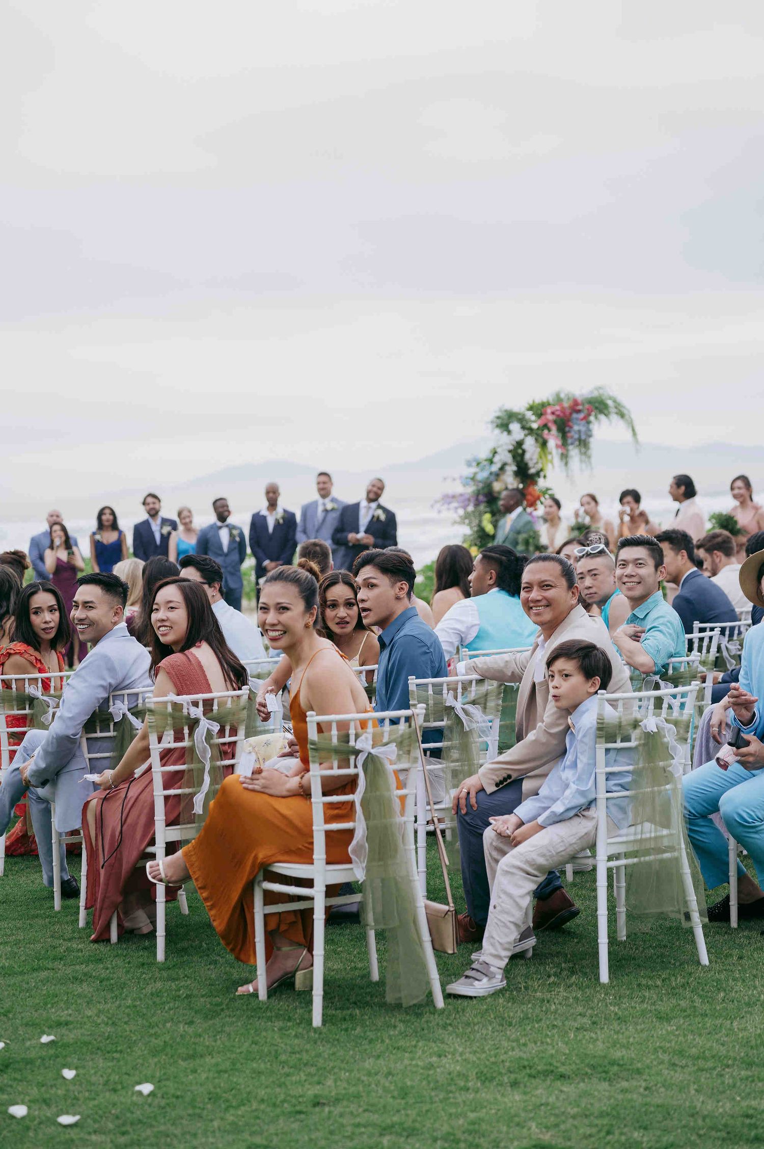 Guests seated in white chairs at an outdoor wedding ceremony with ocean views and floral arch backdrop.