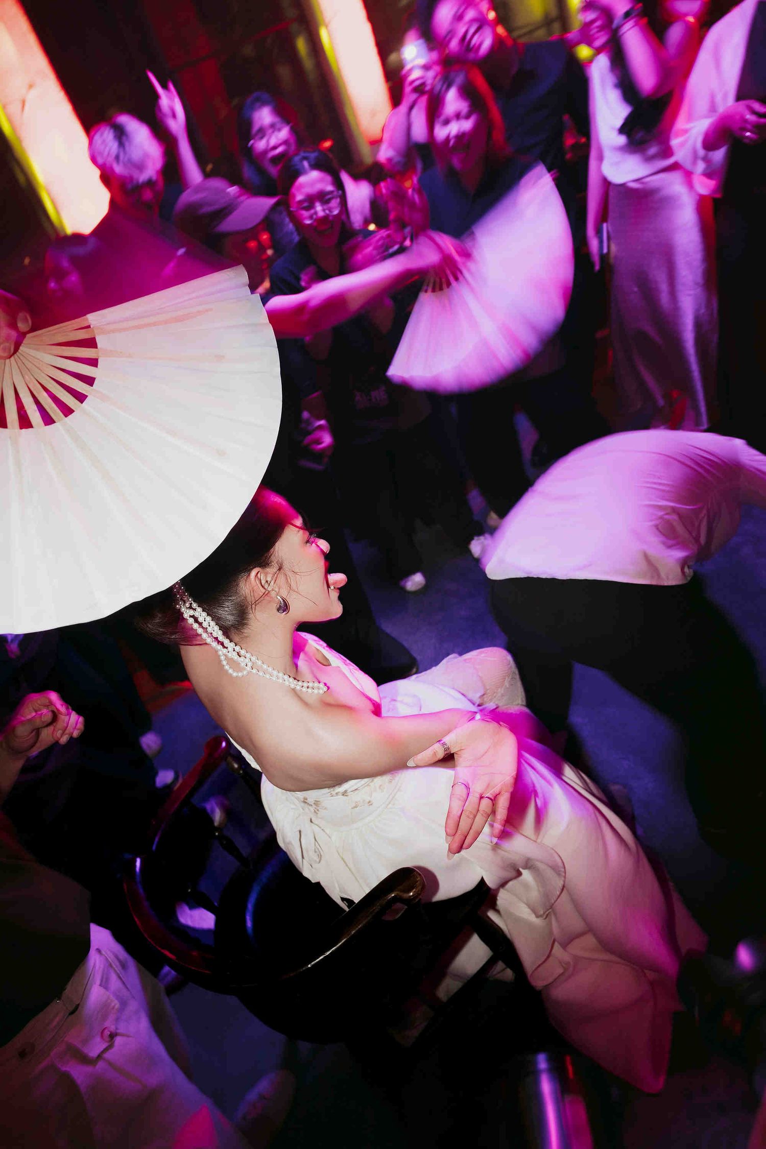 Bride in white dress held aloft by guests waving large white fans at a vibrant wedding reception.