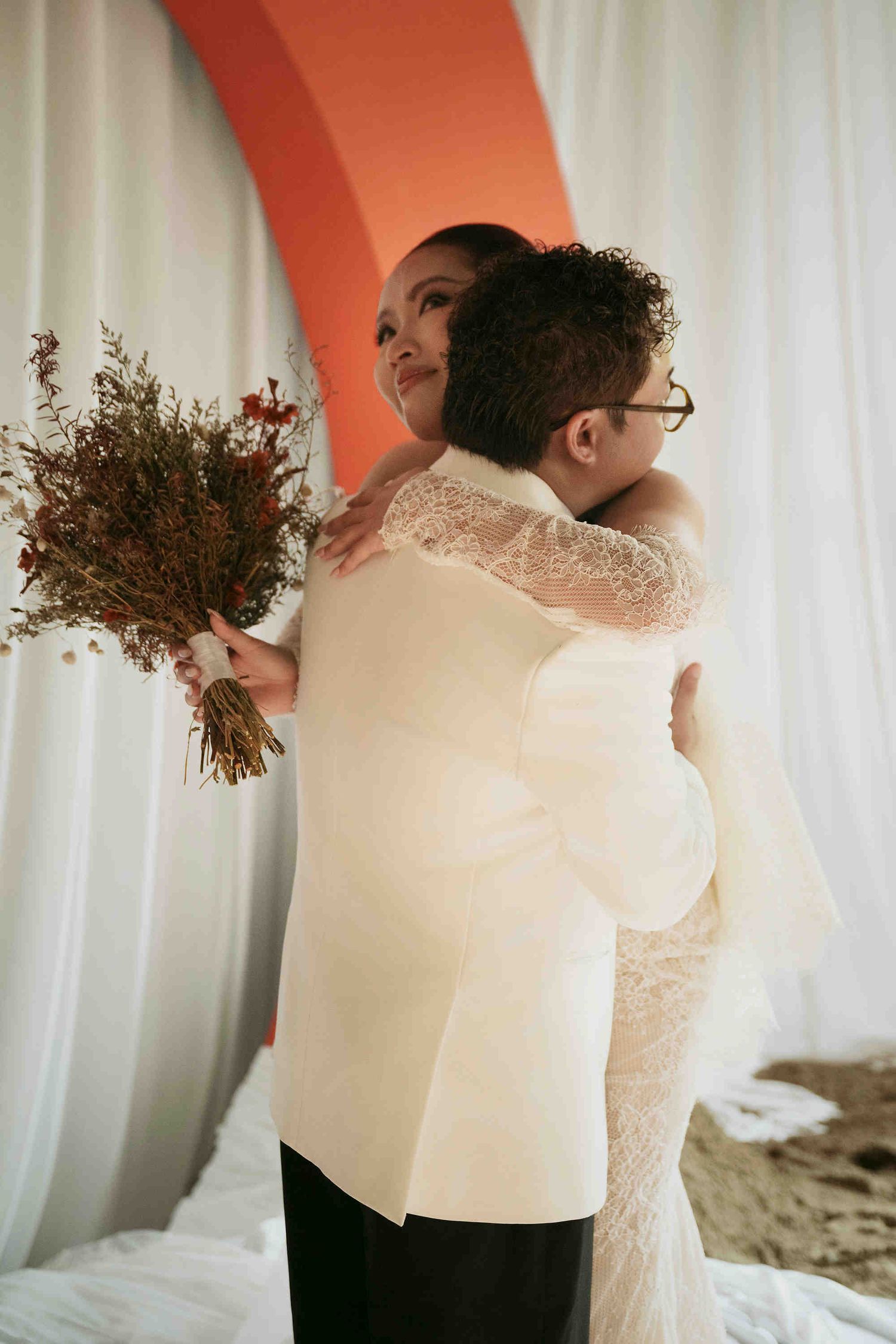 Two brides embracing at wedding ceremony, one in lace gown holding bouquet, other in white blazer, orange arch backdrop.