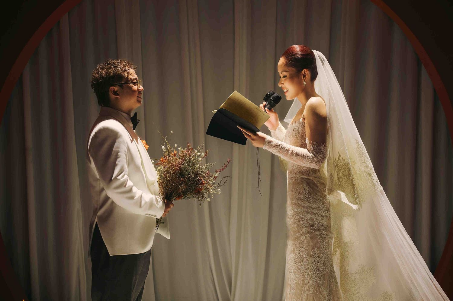 Bride in lace gown reads vows from book while groom holds dried flowers at elegant wedding ceremony.