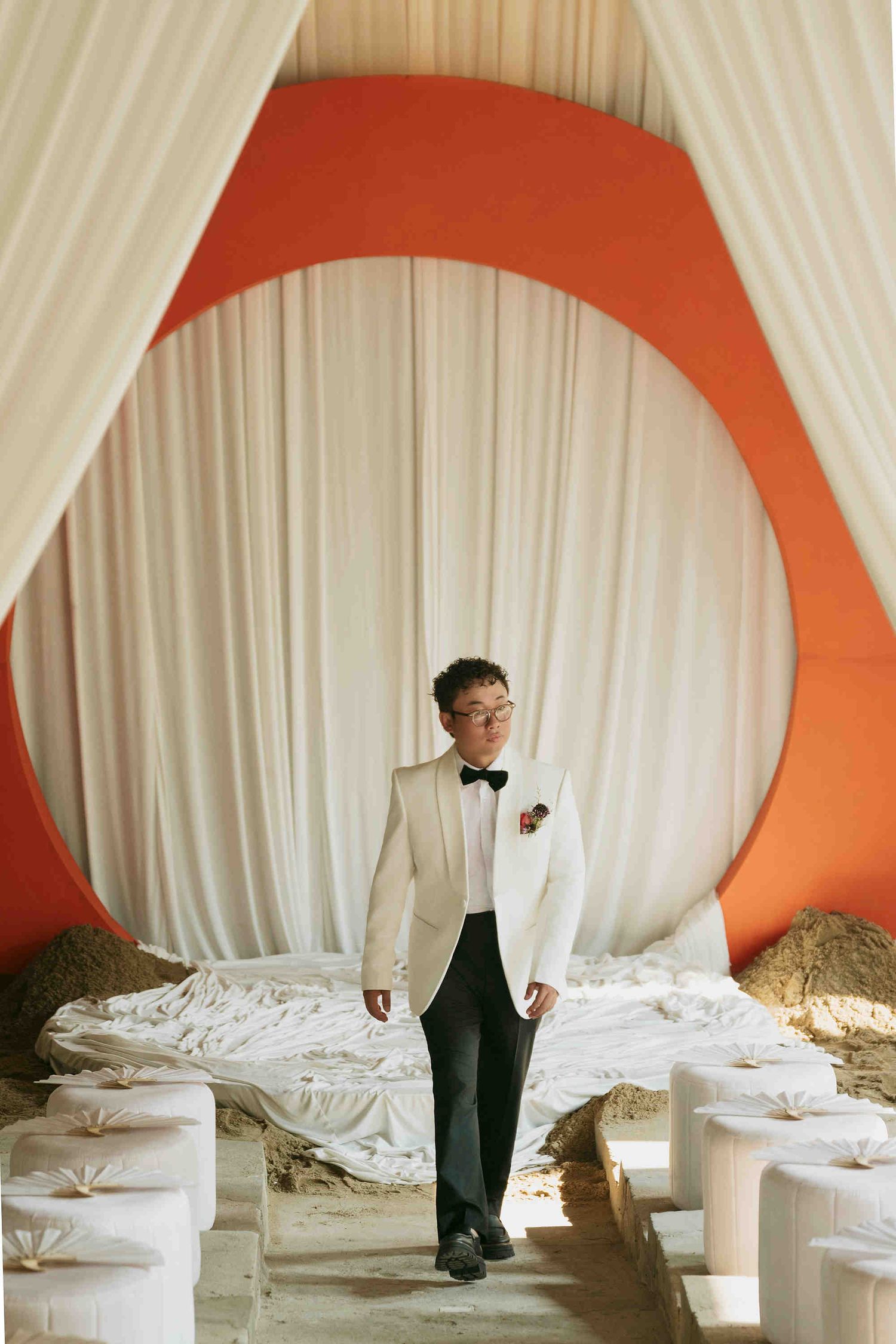 Groom in white tuxedo jacket and black bow tie walks through elegant draped ceremony aisle with orange circular backdrop.