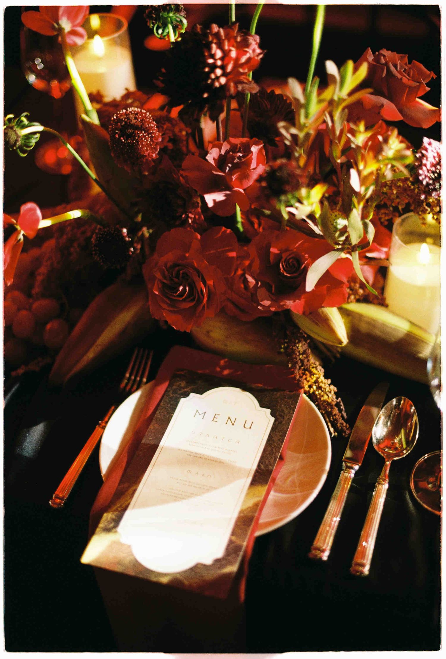 Elegant wedding table setting with deep red roses, candles, and a menu card on a dark romantic tablescape.