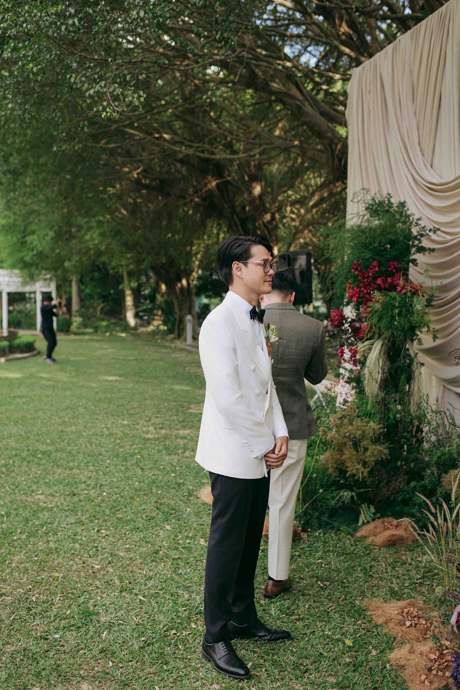 Groom in white tuxedo jacket and black pants stands outdoors at elegant garden wedding ceremony with floral backdrop.