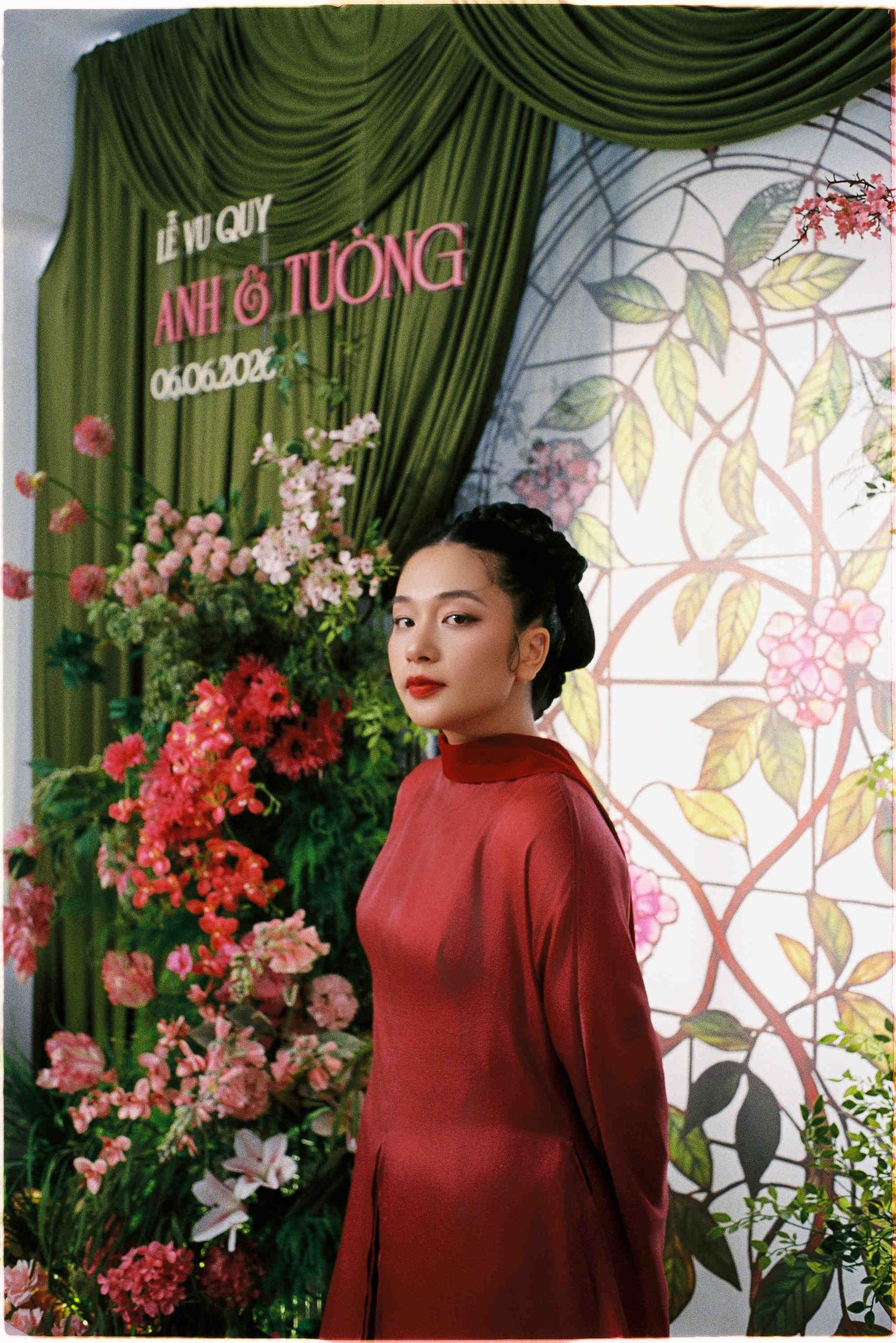 Woman in red ao dai dress poses at a Vietnamese wedding ceremony backdrop with floral decorations.