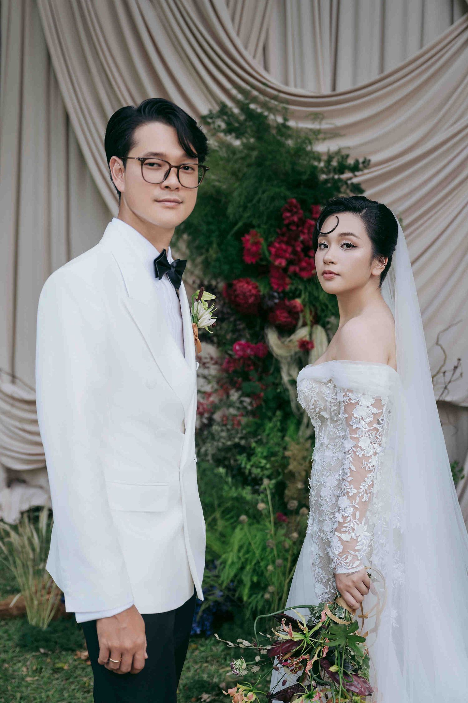 Elegant Asian couple posing at wedding ceremony, groom in white tuxedo and bride in off-shoulder lace gown with veil.
