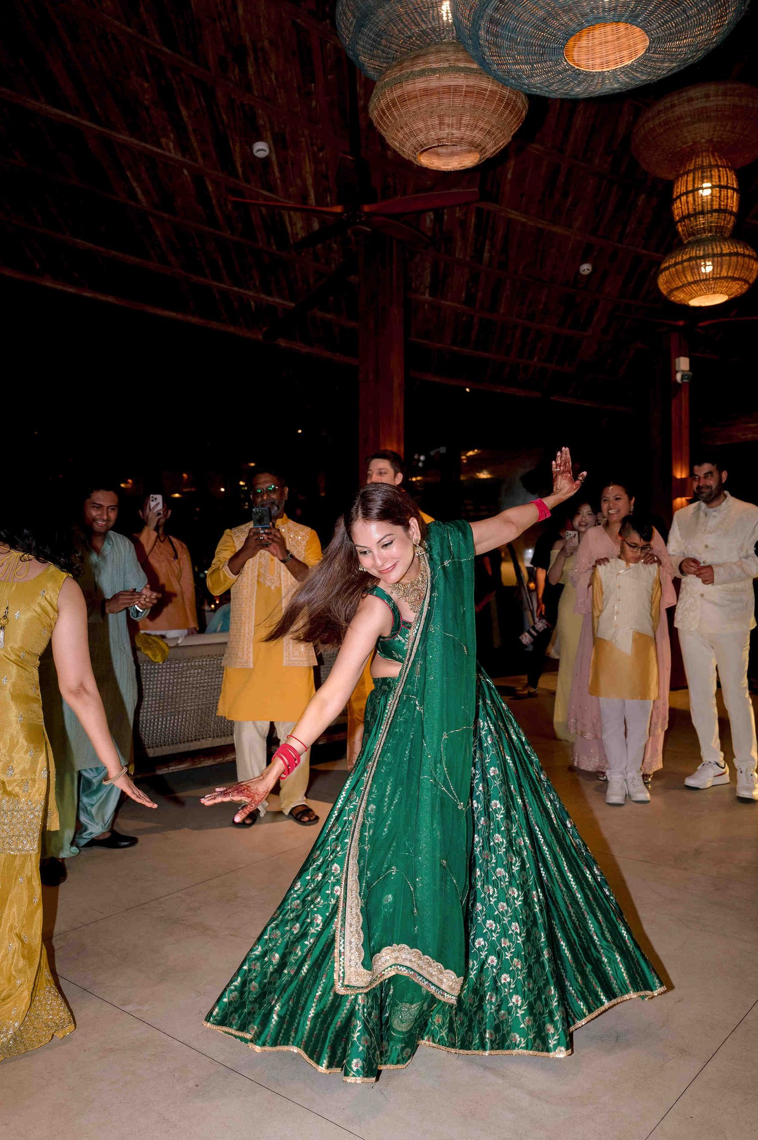 Woman in green lehenga dancing at Indian wedding celebration under wicker pendant lights.