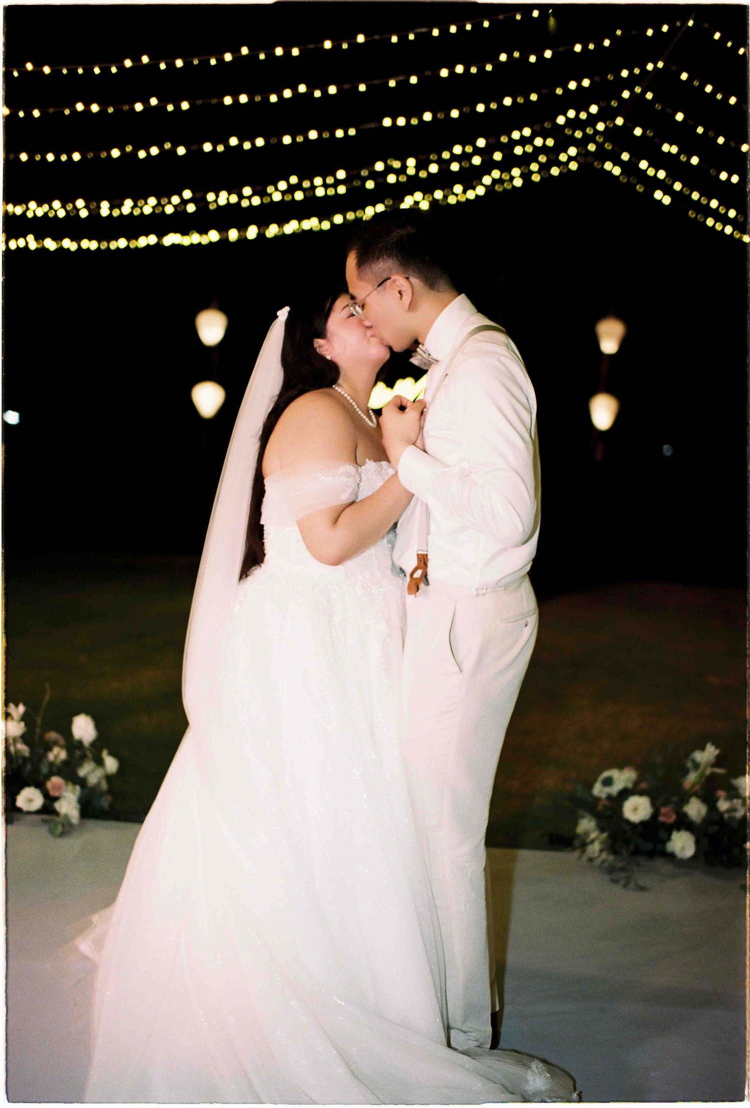 Bride and groom sharing a romantic first dance kiss under twinkling fairy lights at an outdoor evening wedding reception.