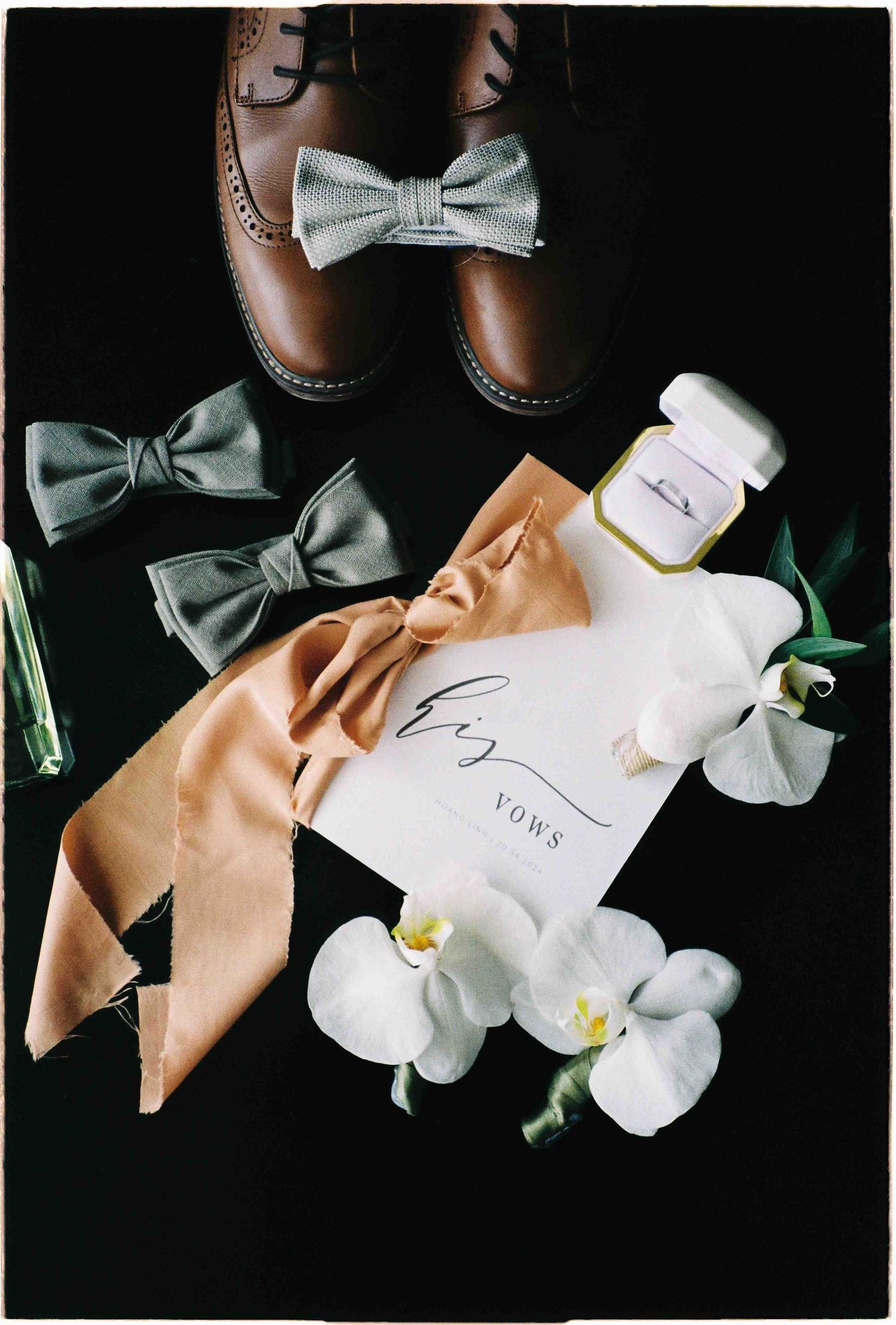 Wedding flat lay with brown leather shoes, gray bow ties, vows card, ring box, orchids, and a peach satin ribbon.