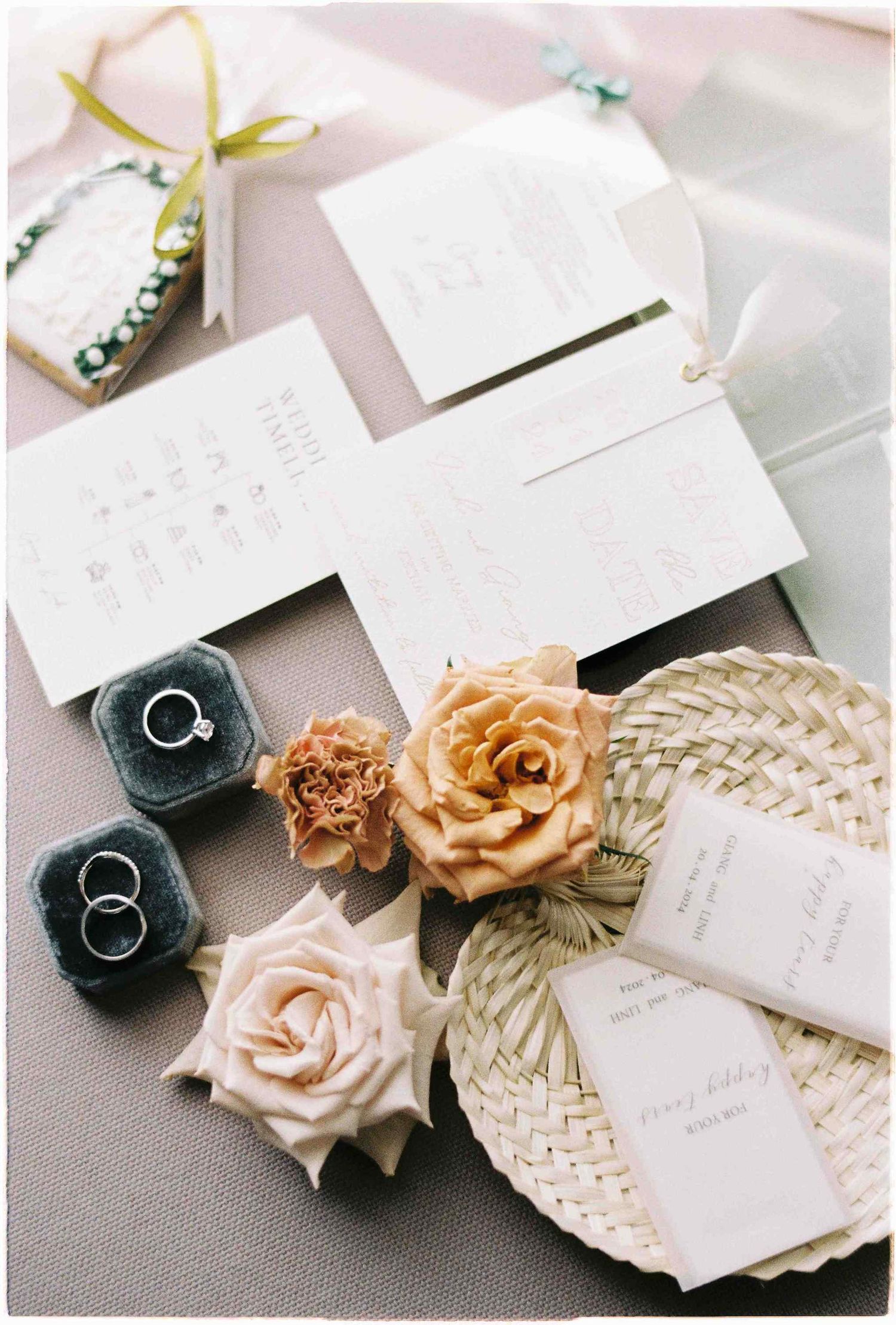 Elegant wedding flat lay with invitation suite, rings in velvet boxes, and floral accents on a soft lavender surface.