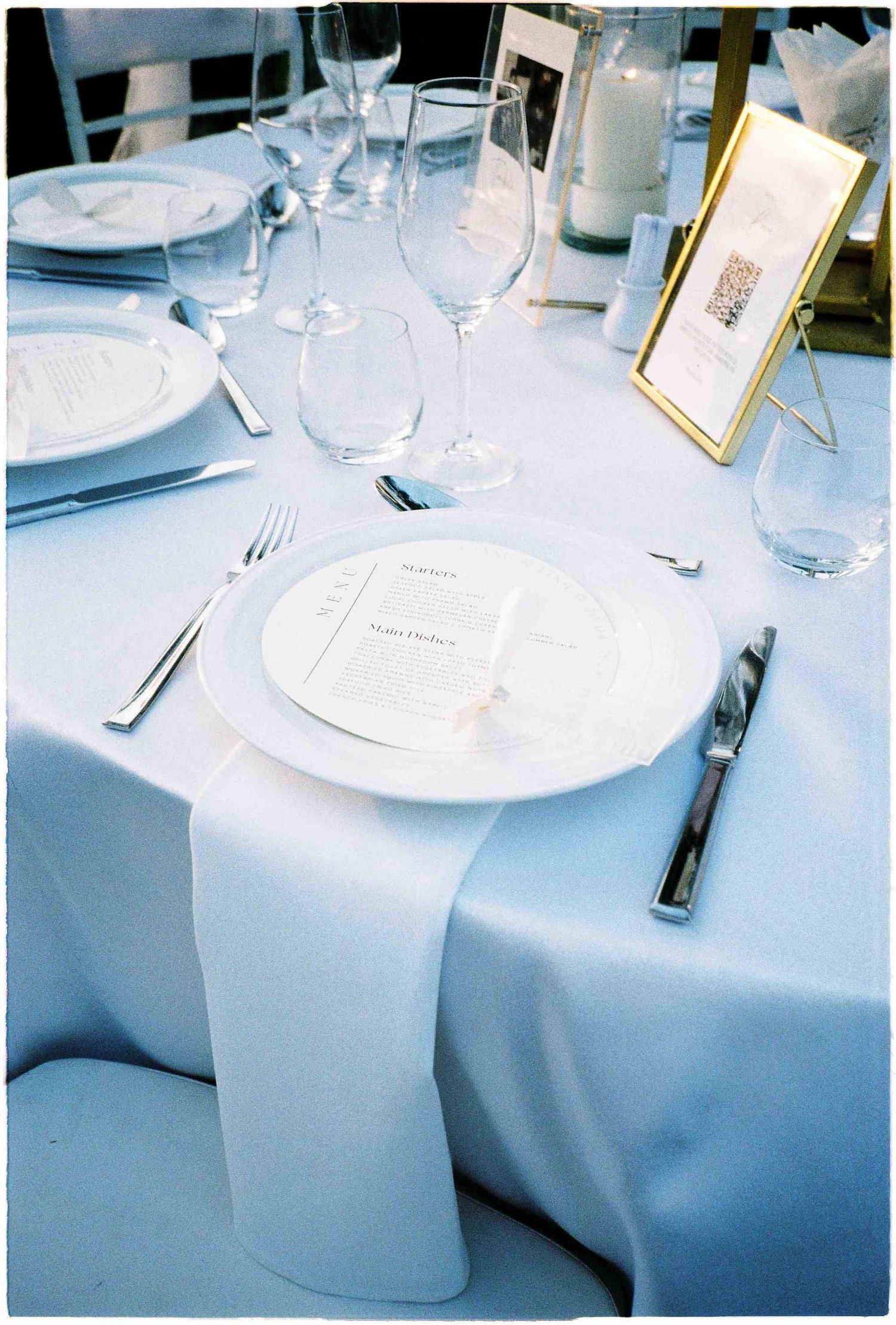 Elegant white wedding table setting with fine china, crystal glasses, silverware, and a gold-framed menu card.