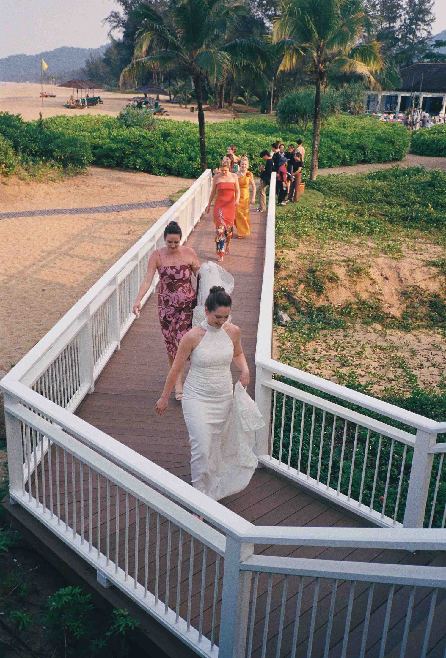 Lang Co wedding: wedding party walking down white-railed wooden boardwalk with palm trees and beach in background, by AnhVuWedding.