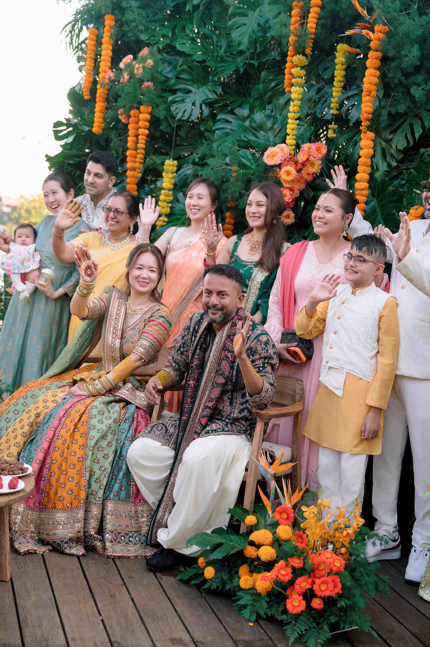 Joyful Indian wedding celebration with couple seated under marigold garlands, surrounded by smiling guests in colorful traditional attire.