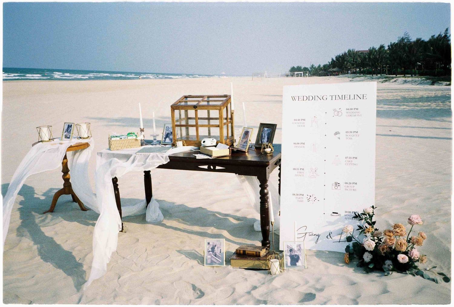 Beach wedding timeline display table with floral arrangements, candles, and decor set on white sand by the ocean.