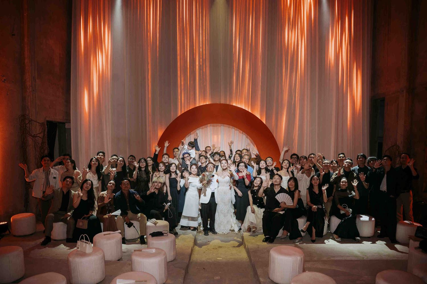 Large wedding group photo with bride and groom surrounded by guests in elegant venue with warm orange lighting.