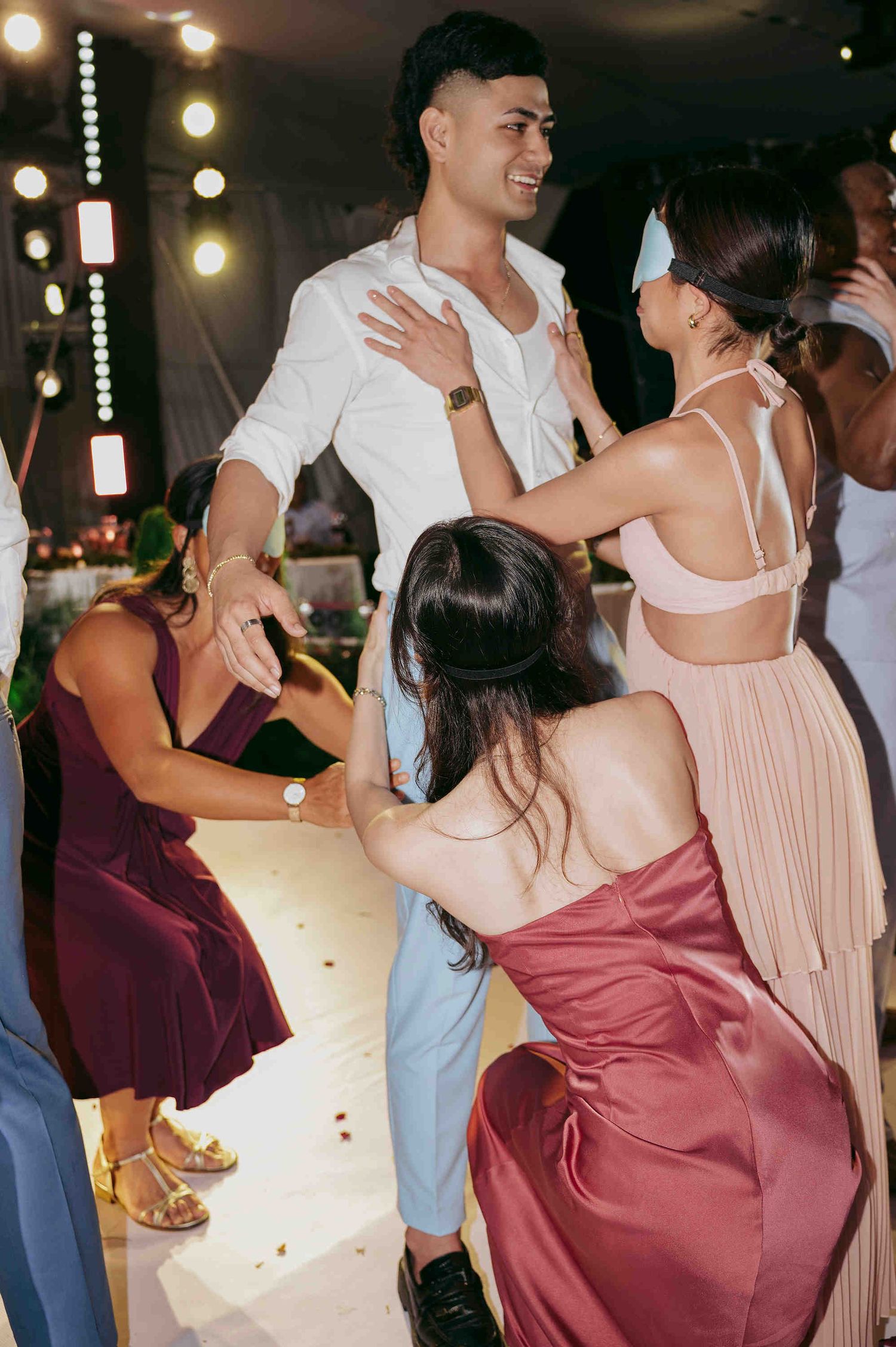 Guests dancing together at a wedding reception, wearing formal attire in burgundy, pink, and white under warm string lights.