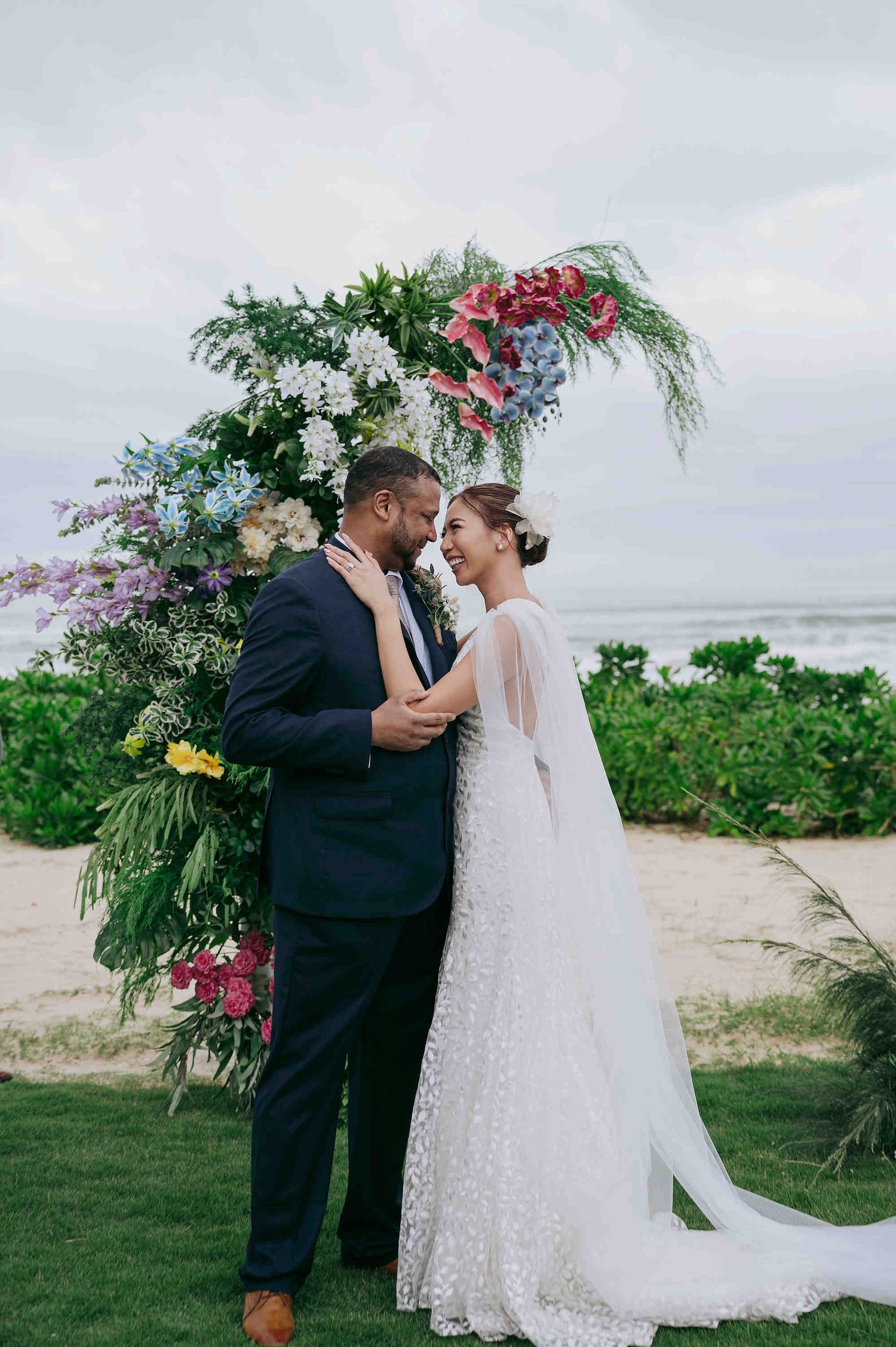 Couple sharing a romantic moment at a beachside wedding ceremony under a colorful floral arch with ocean views.