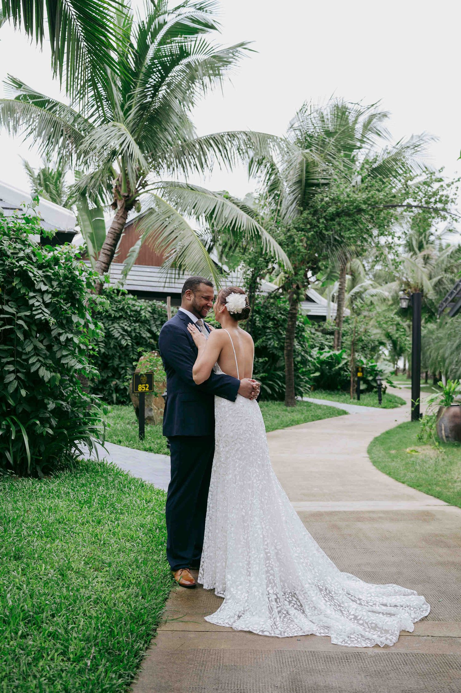 A bride and groom embrace on a tropical garden pathway surrounded by lush palm trees in their wedding attire.