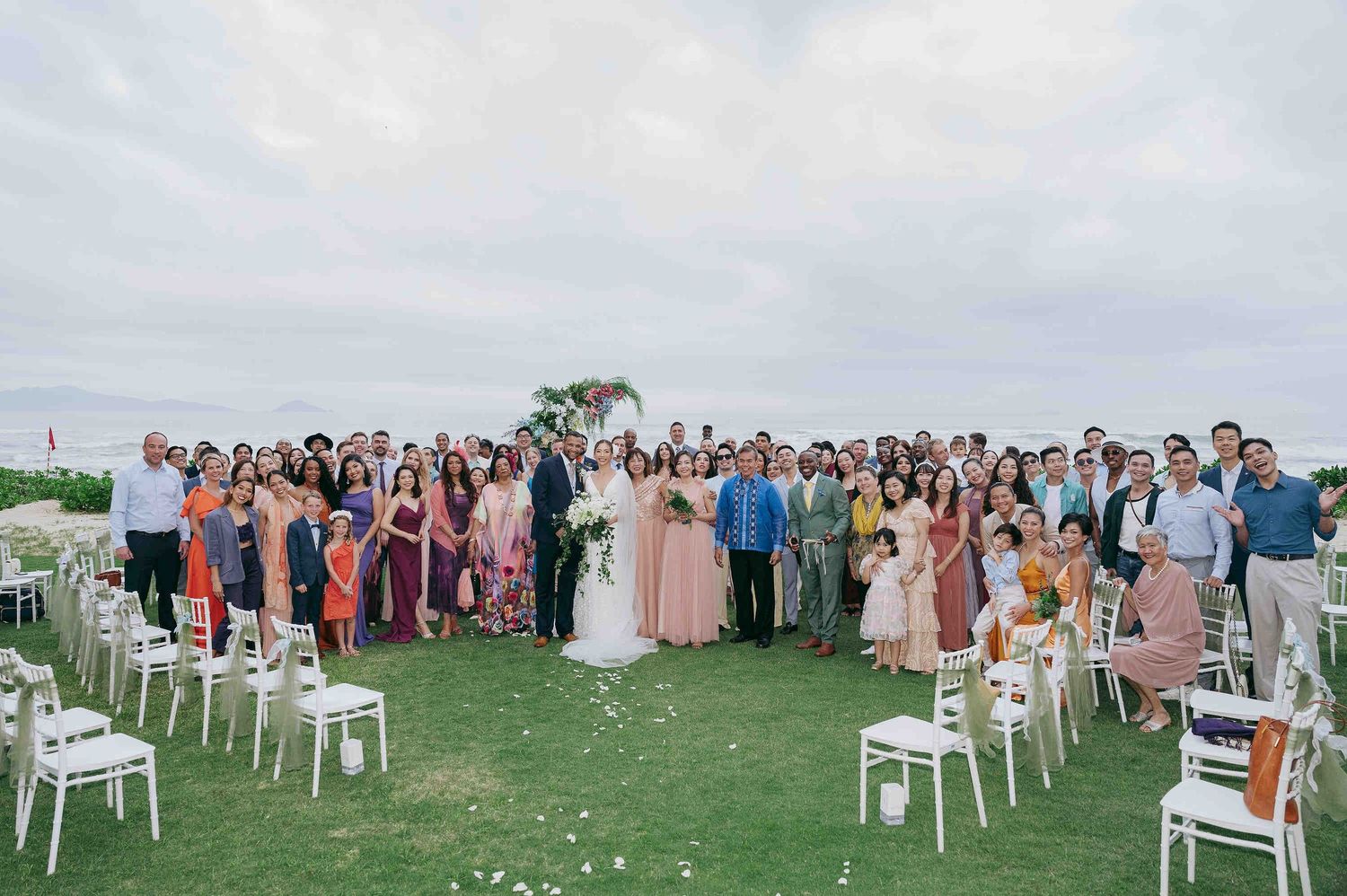 Large wedding group photo with bride and groom surrounded by guests on a green lawn at an outdoor seaside ceremony.