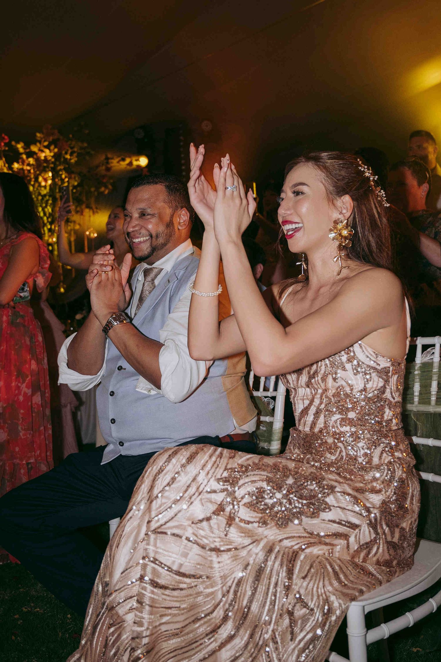 Couple in elegant attire clapping joyfully at a wedding reception, woman in gold sequin gown with floral hair accessory.