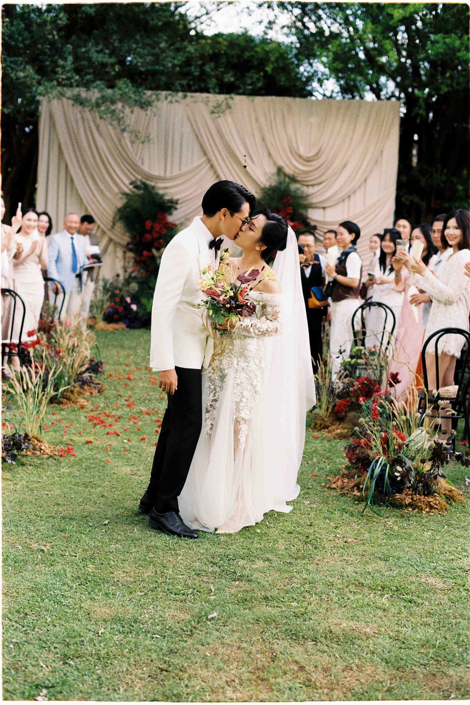 Bride and groom share first kiss at outdoor wedding ceremony with floral arrangements and draped backdrop.