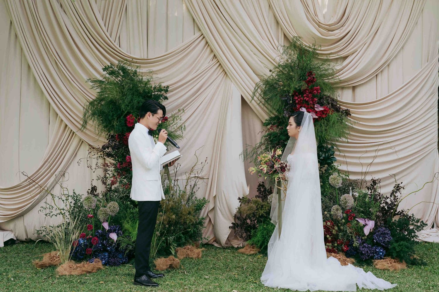Couple exchanging vows at an elegant outdoor wedding ceremony with lush floral arrangements and draped fabric backdrop.