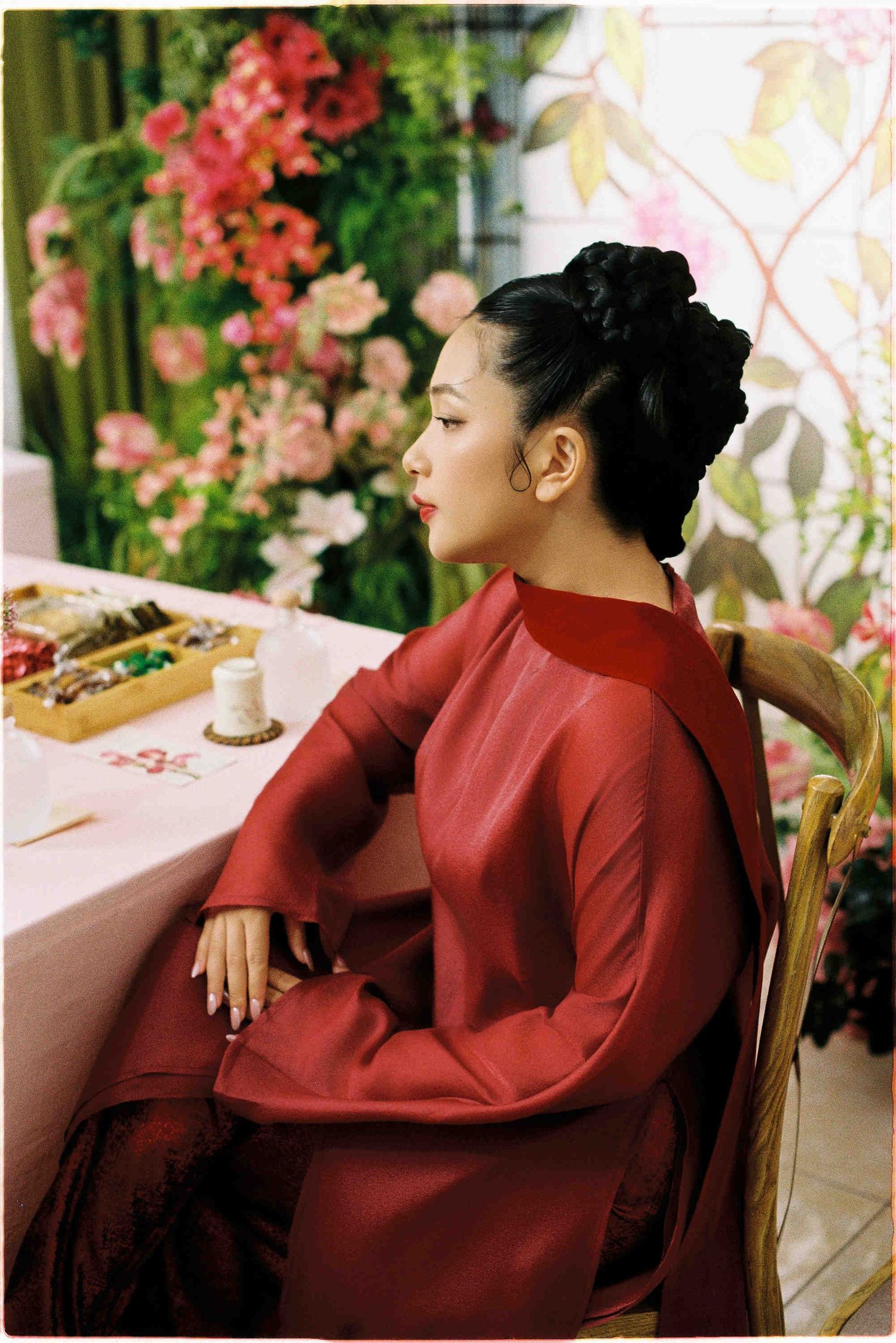 Woman in red áo dài sits gracefully at a floral table, hair in elegant updo, surrounded by pink blooms.
