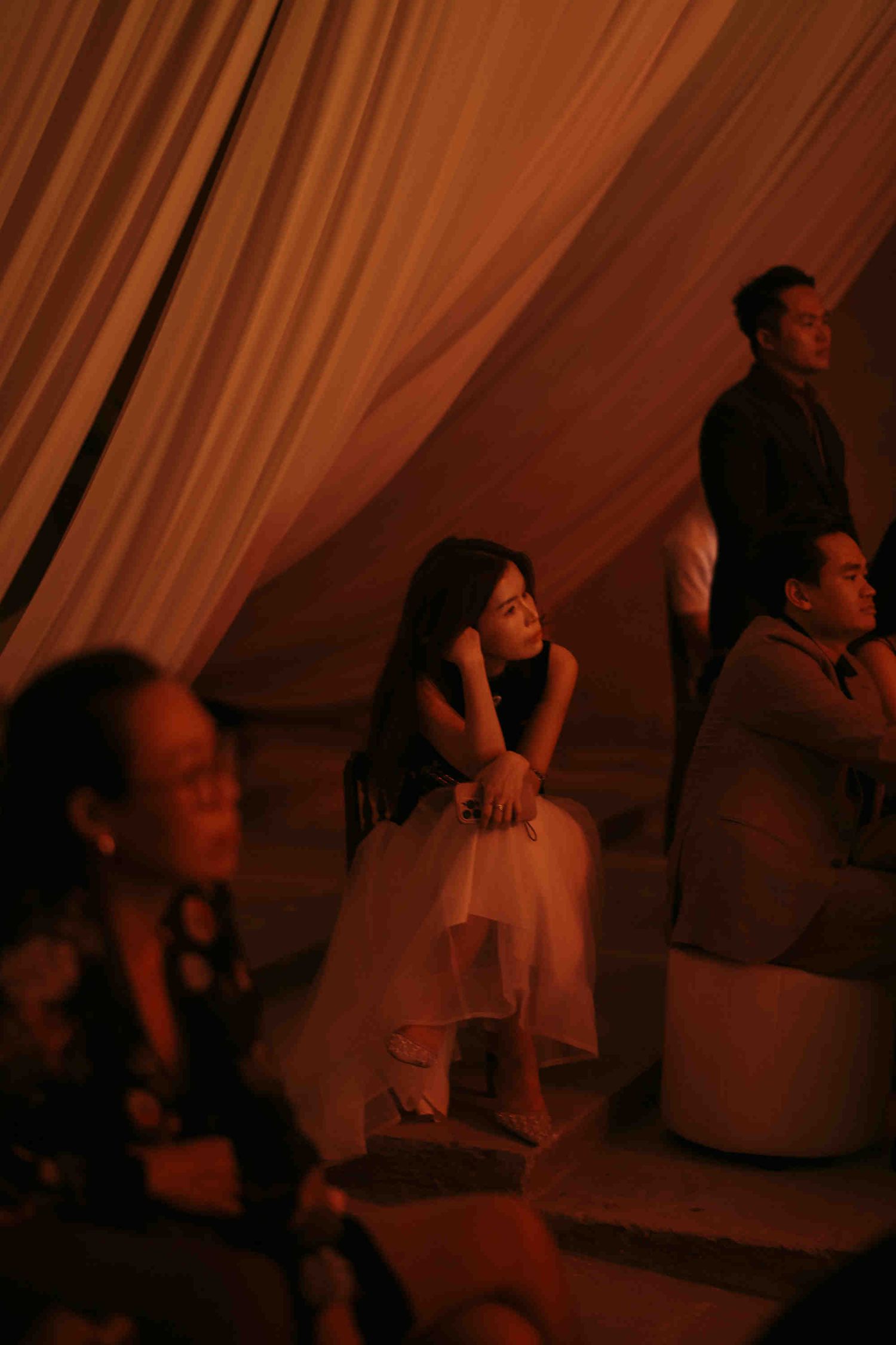 Woman in white skirt sits pensively at an elegant evening event under warm amber draped fabric lighting.