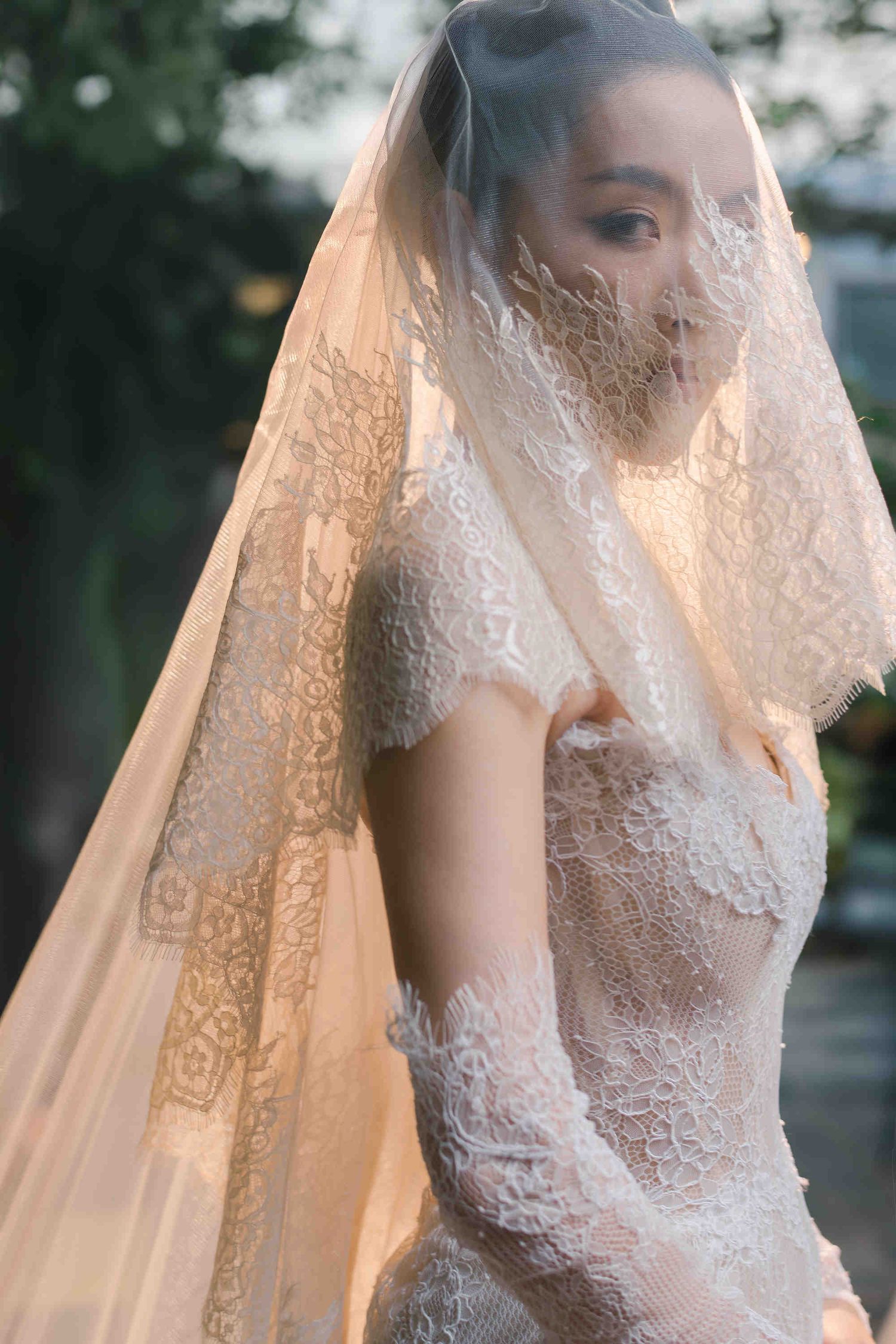 Bride in elegant lace wedding dress and veil draped over face, standing outdoors in soft natural light.