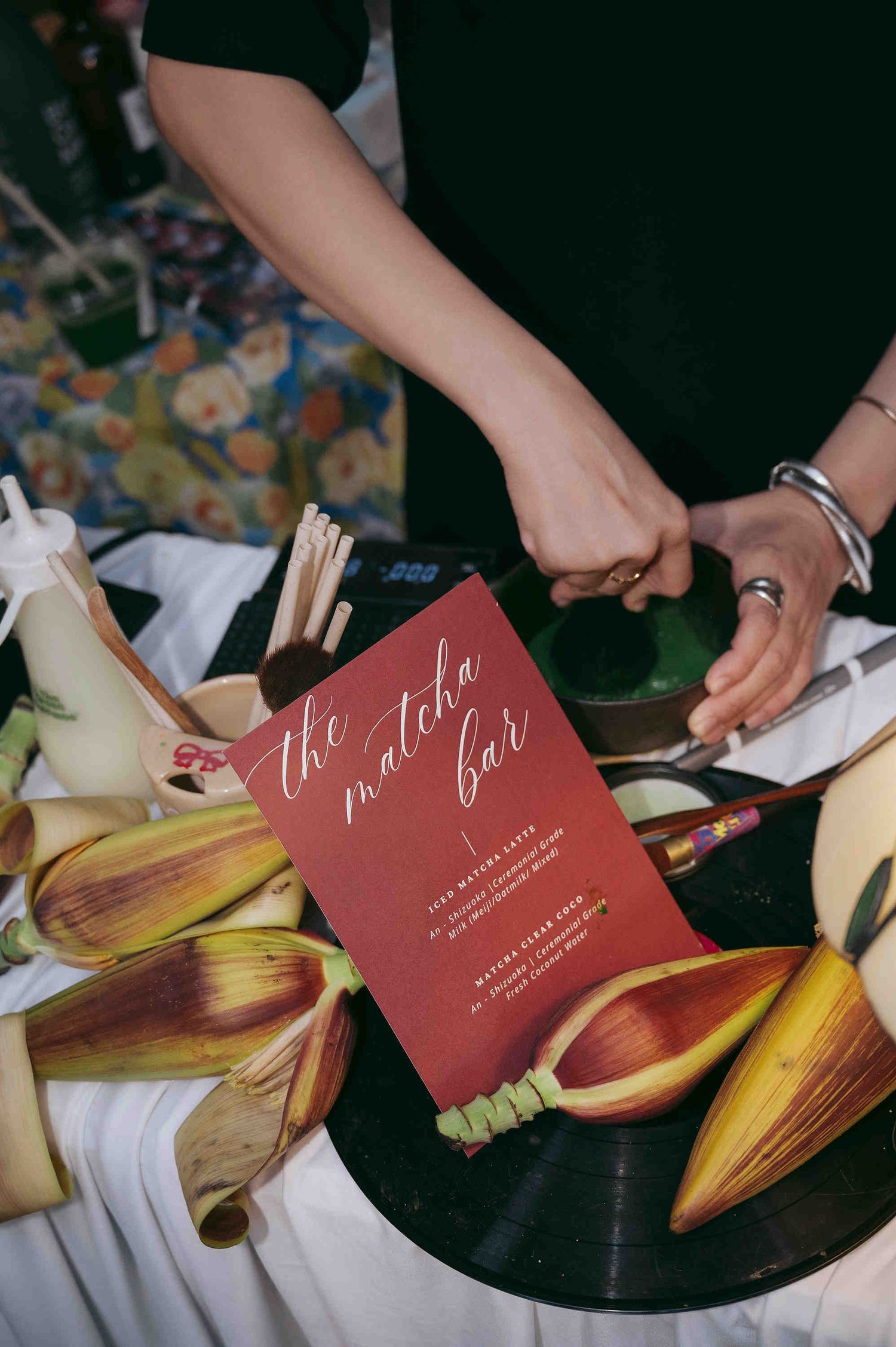 A red matcha bar menu card displayed on a table with tropical leaves, vinyl record, and matcha preparation tools.