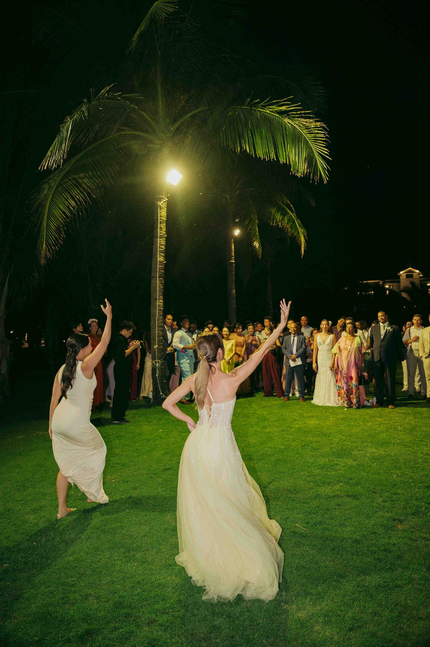 Bride throws bouquet as guest leaps to catch it during outdoor tropical wedding reception at night.