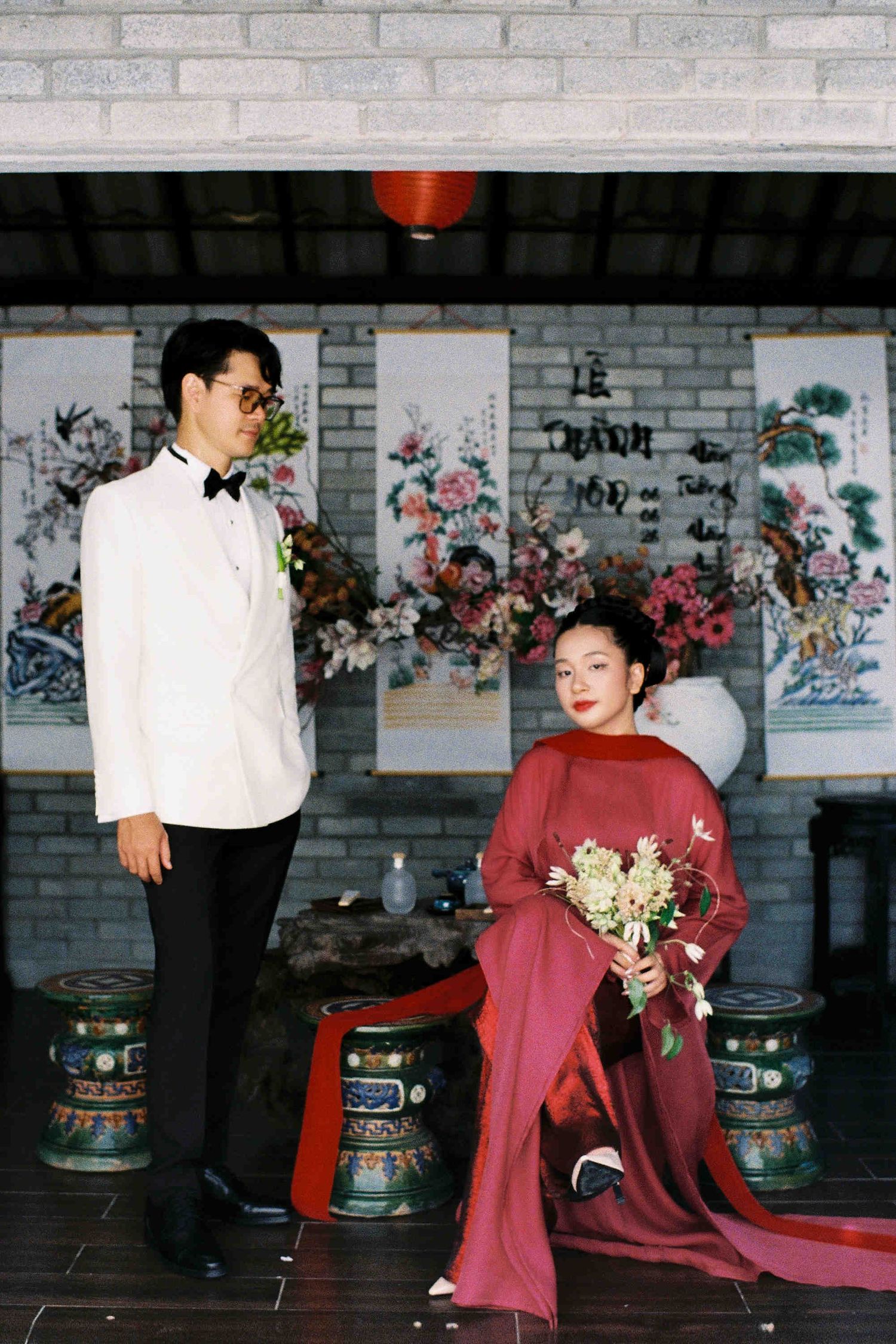 Vietnamese couple in traditional wedding attire, groom in white tuxedo, bride in red ao dai holding bouquet in ornate setting.