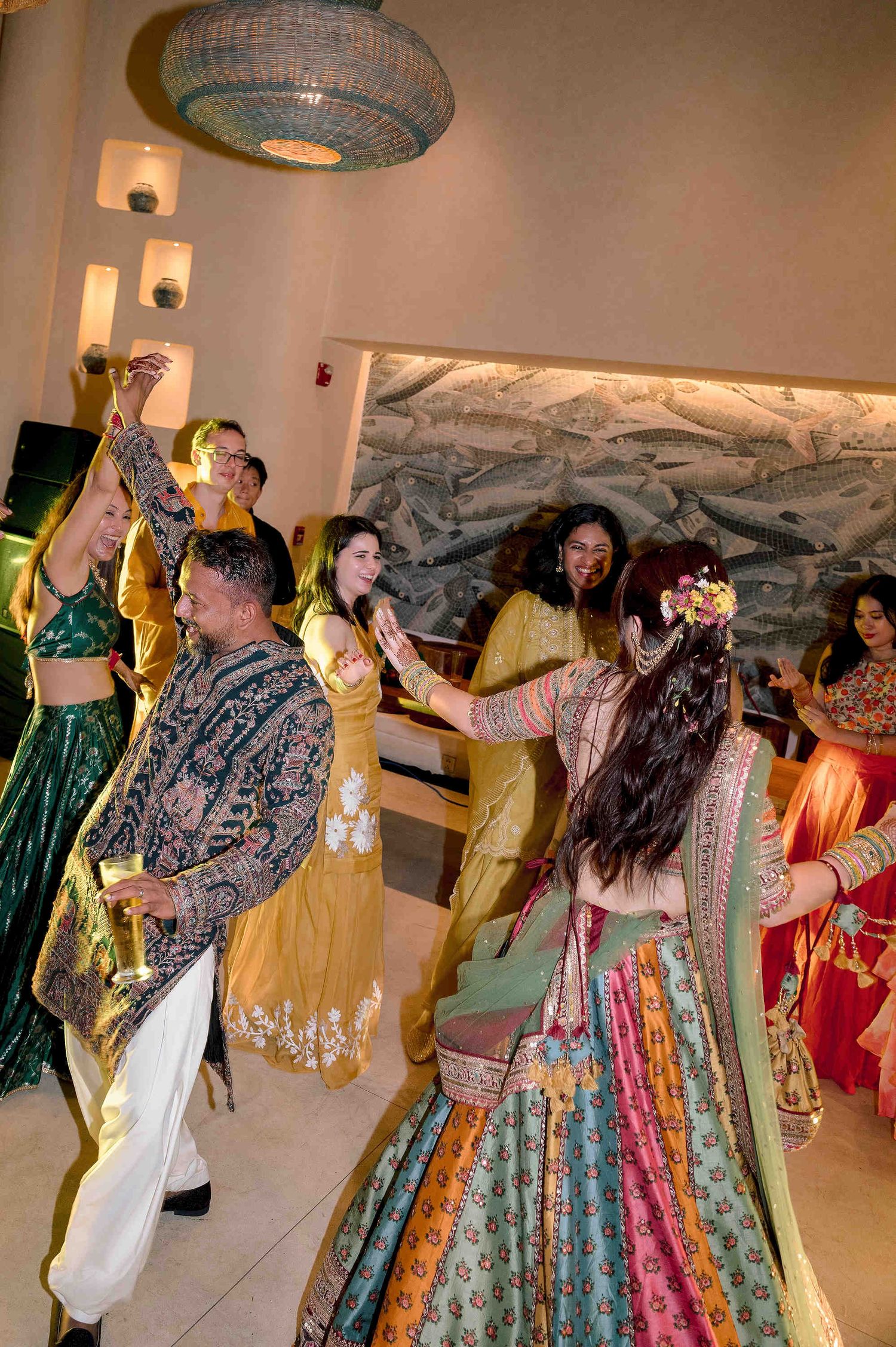 Guests in vibrant traditional Indian attire dance joyfully at a colorful wedding celebration indoors.